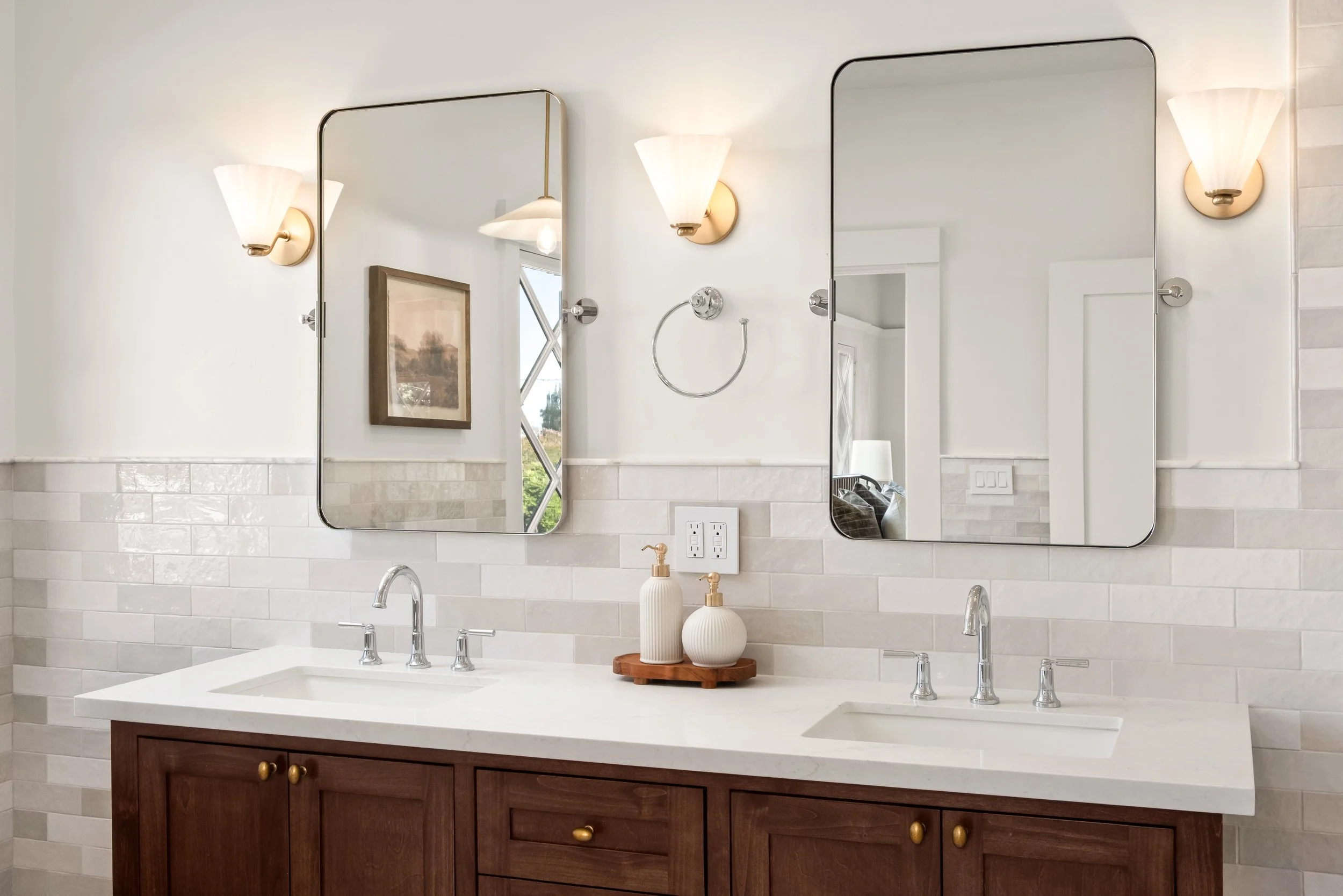 Bathroom vanity with double sinks, two mirrors, wall sconces, a framed painting, and decorative vases on a wooden tray.