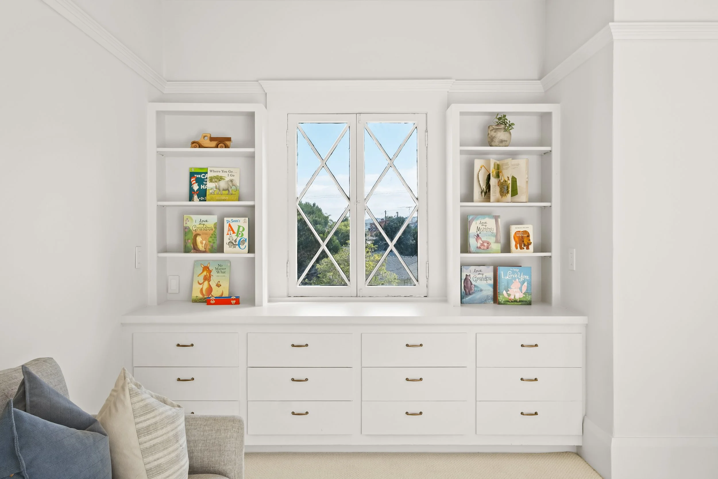 White built-in bookshelf with books and decorative items positioned in front of a window, with a corner of a light-colored sofa and pillows visible in the lower left corner.