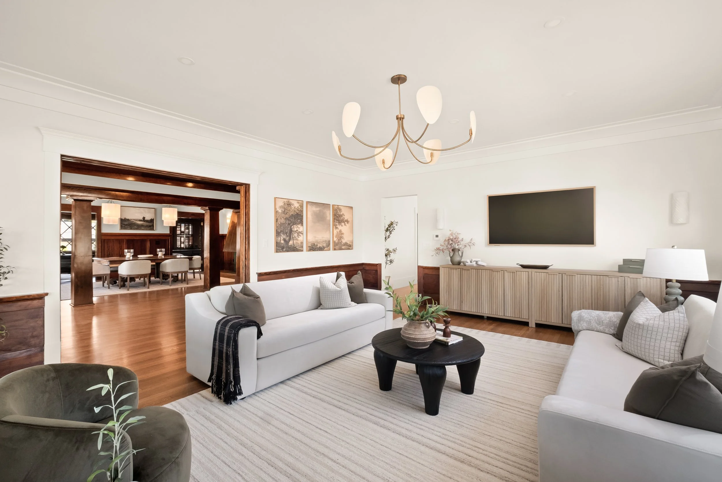 Living room with white sofas, a black coffee table, a wall-mounted flat-screen TV, framed artwork, potted plant, and modern lighting fixtures.