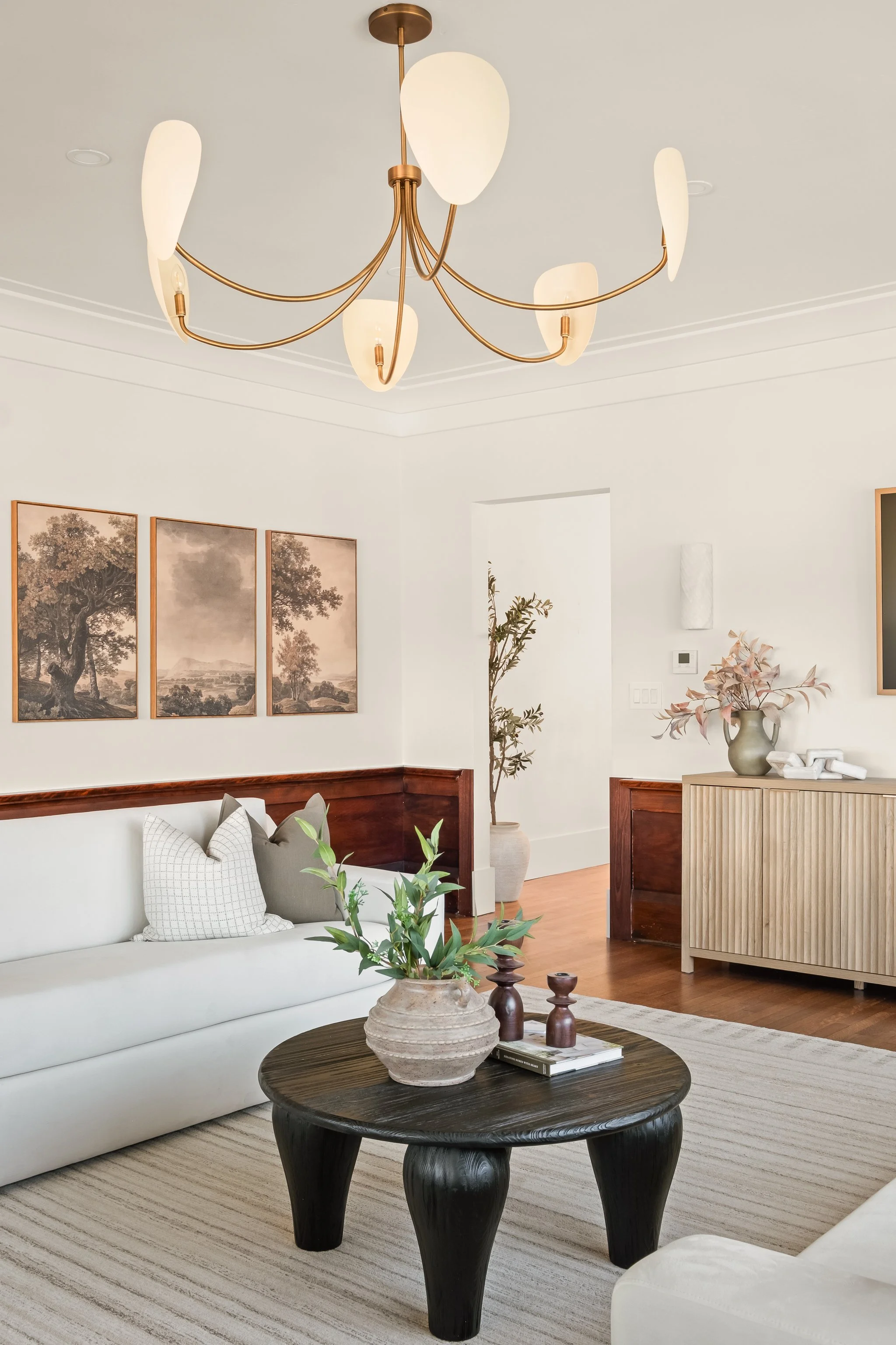 Living room with a white sofa, neutral pillows, wooden flooring, a black coffee table with decorative items, a beige striped rug, and a chandelier hanging from the ceiling. Wall art features black-and-white trees.