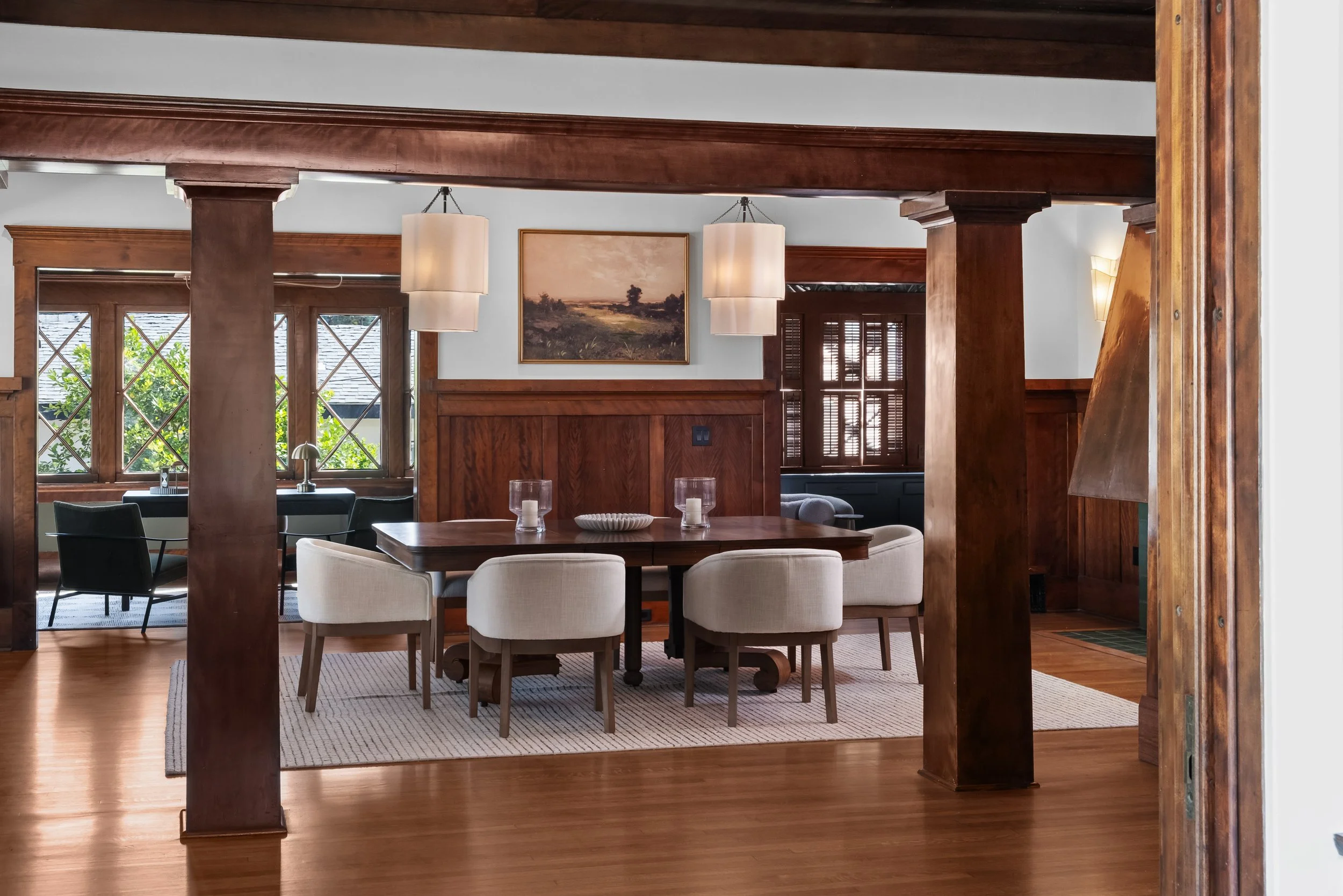 A dining room with a wooden ceiling and beams, white walls, large windows with diamond-shaped panes, and a dining table surrounded by white and black chairs. There is a framed landscape painting on the wall and curtains on the windows. Two hanging pe