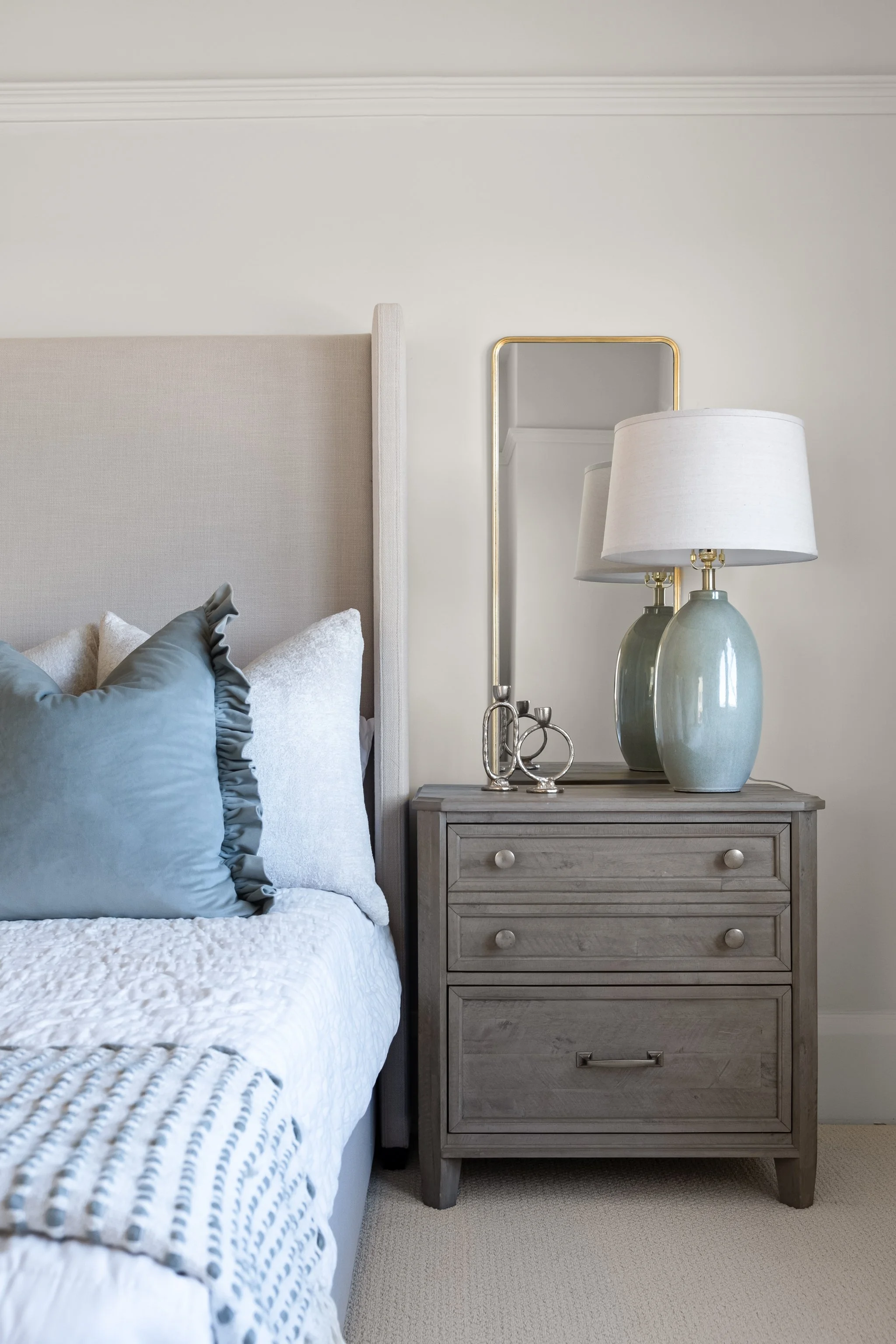 A bedroom nightstand with two green vases and a table lamp, next to a bed with white and blue pillows and a white quilt, against a light-colored wall with decorative molding.
