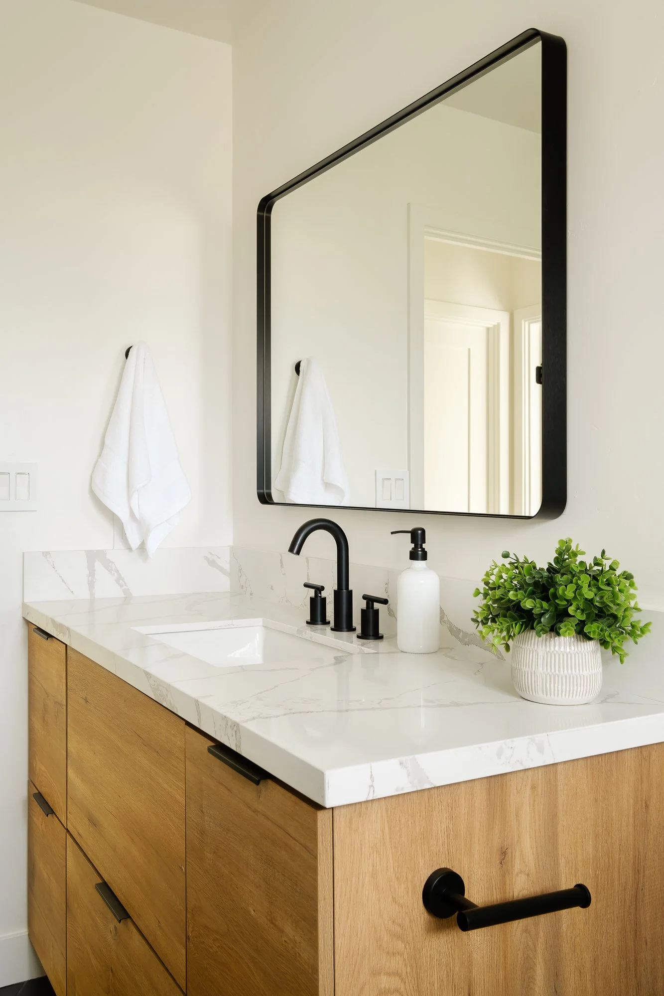 Modern bathroom vanity with a white marble countertop, a black faucet, a wall mirror with black frame, a white soap dispenser, and a potted plant. White towels hanging on hooks are reflected in the mirror.