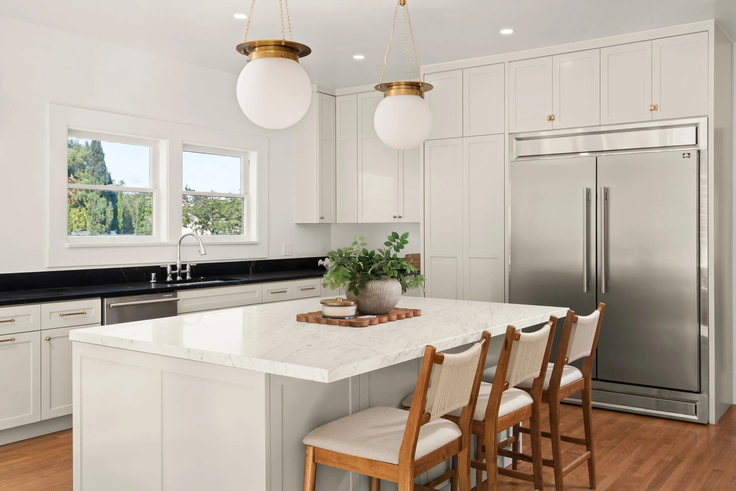 Modern kitchen with white cabinets, black countertop, stainless steel appliances, two pendant lights, a window with greenery outside, a white marble kitchen island, a potted plant, and wooden chairs.