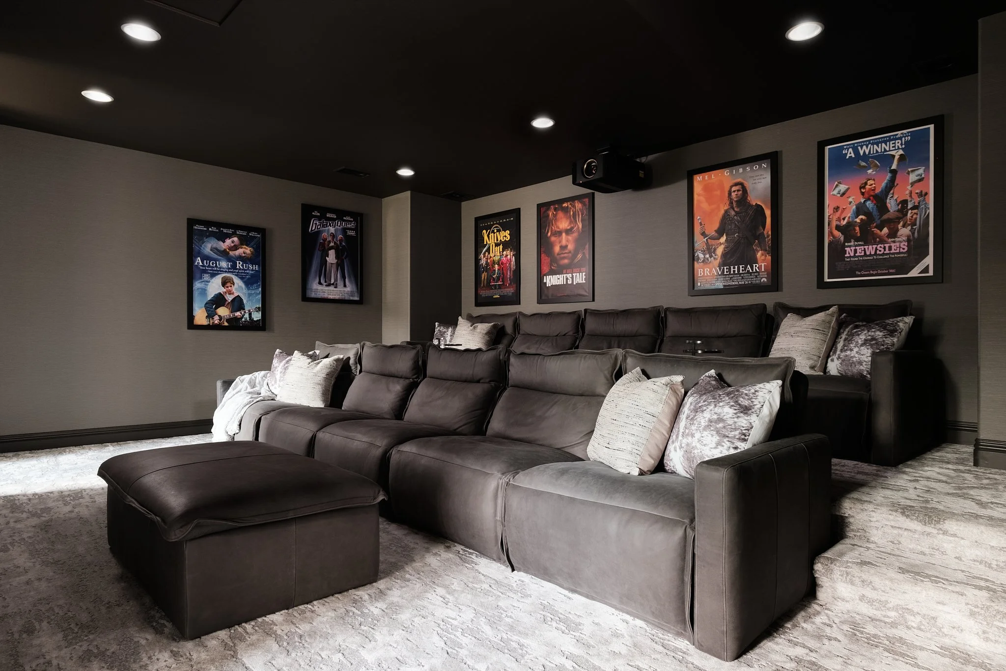 A cozy home theater room with gray plush sectional sofa and pillows. Dark ceiling with recessed lighting. Walls decorated with framed movie posters, including August Rush, Galaxy Quest, Knives Out, Brave Heart, and Newsies.