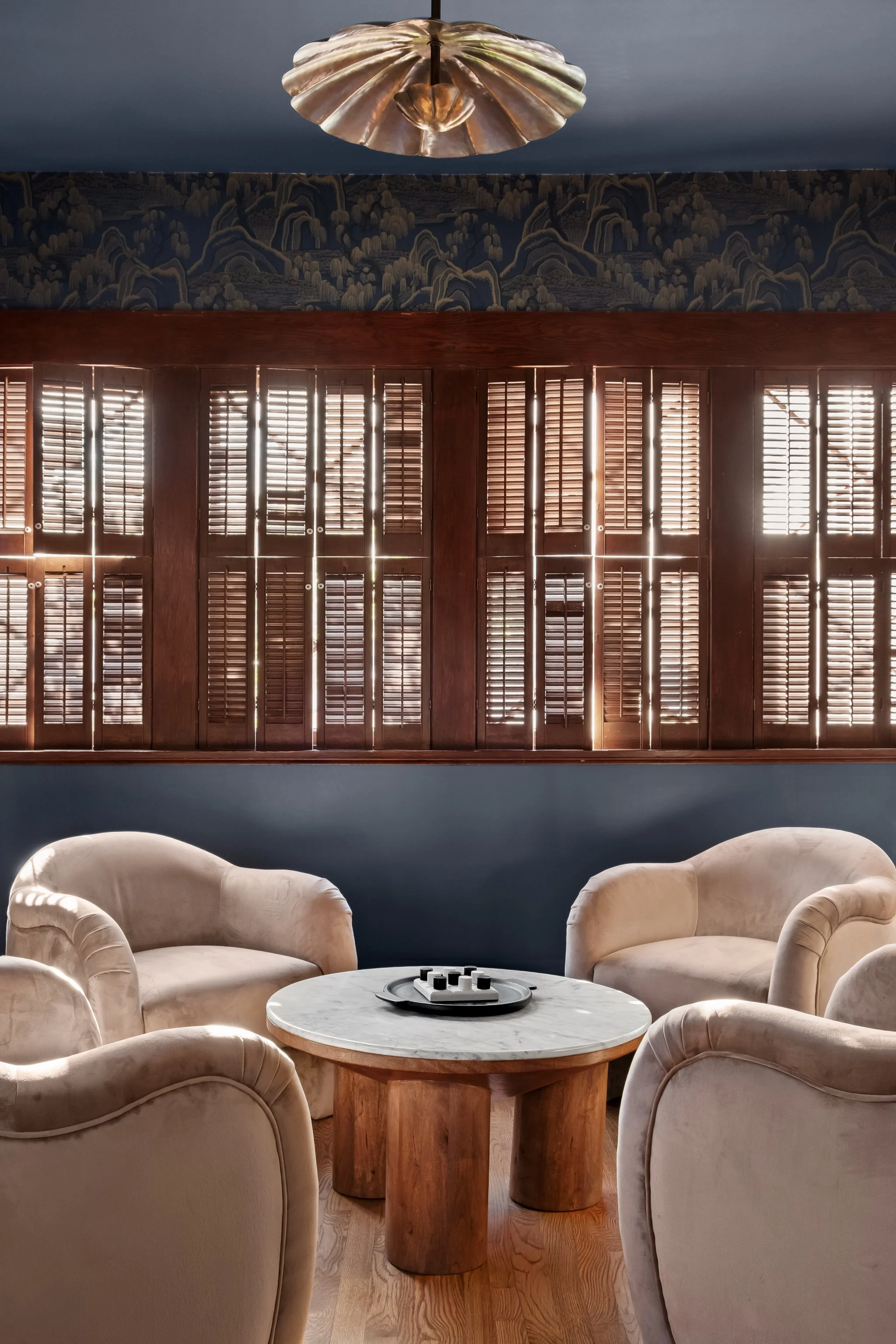 A cozy sitting area with four beige velvet armchairs and a round marble-top coffee table with a decorative tray on it. In the background, a large window with wooden shutters allows sunlight to filter in, and above the window, there's a dark blue wall