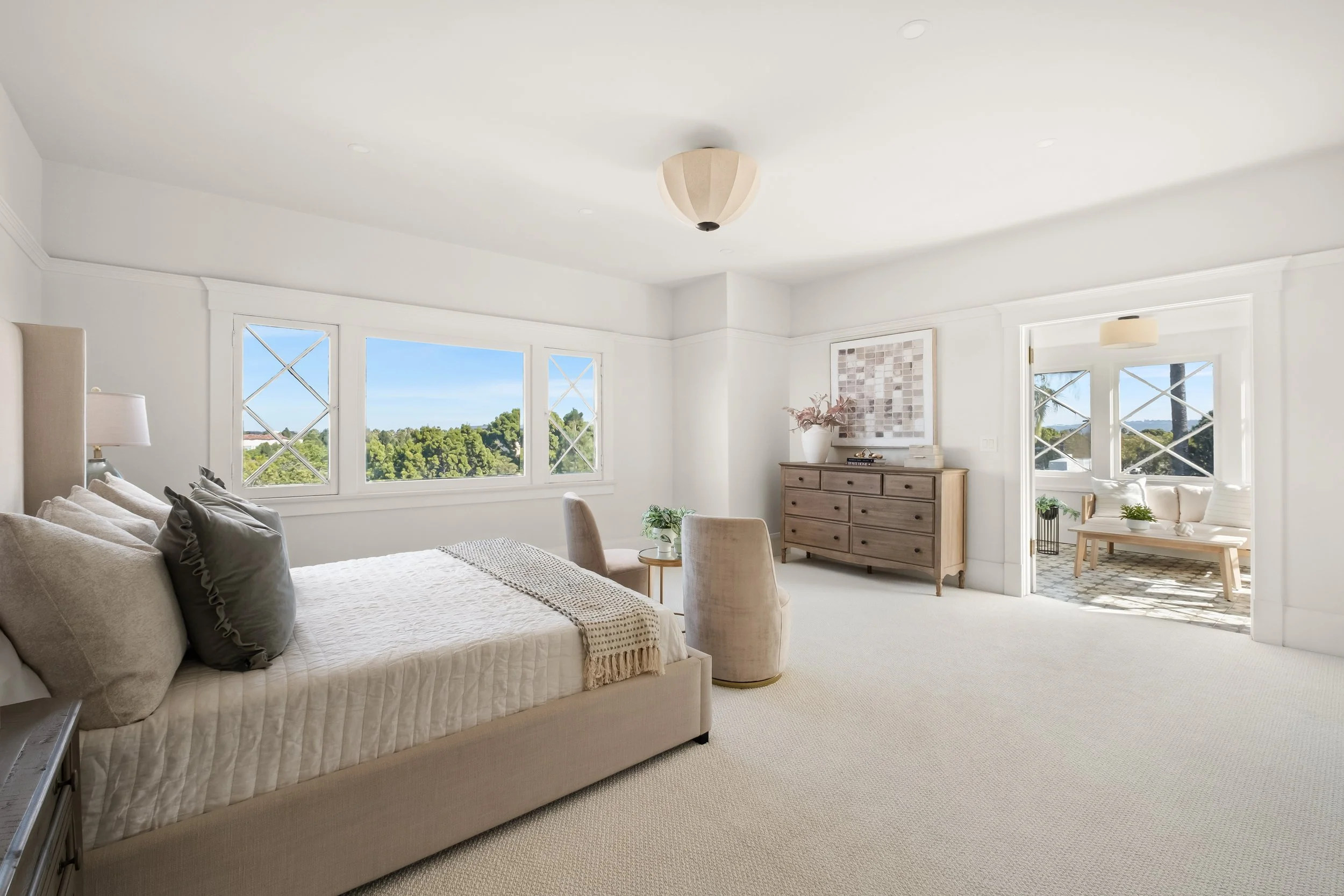 bright living room with large windows, beige sofa with pillows, wooden dresser with decorative items, and an adjoining room with outdoor view