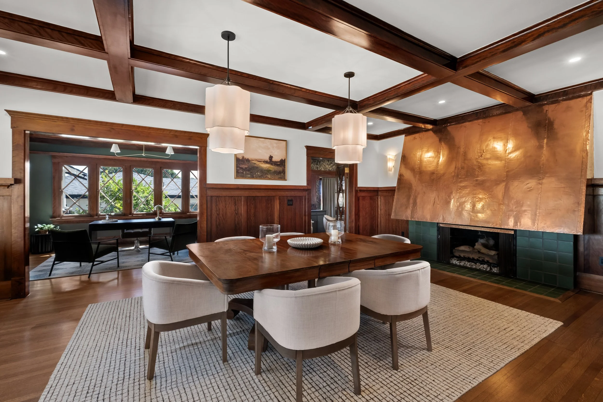 A cozy living and dining room with wood-paneled walls, wooden ceiling beams, and a large copper fireplace. The dining table is surrounded by beige upholstered chairs, and the room features large windows with views of greenery outside. Pendant lights 