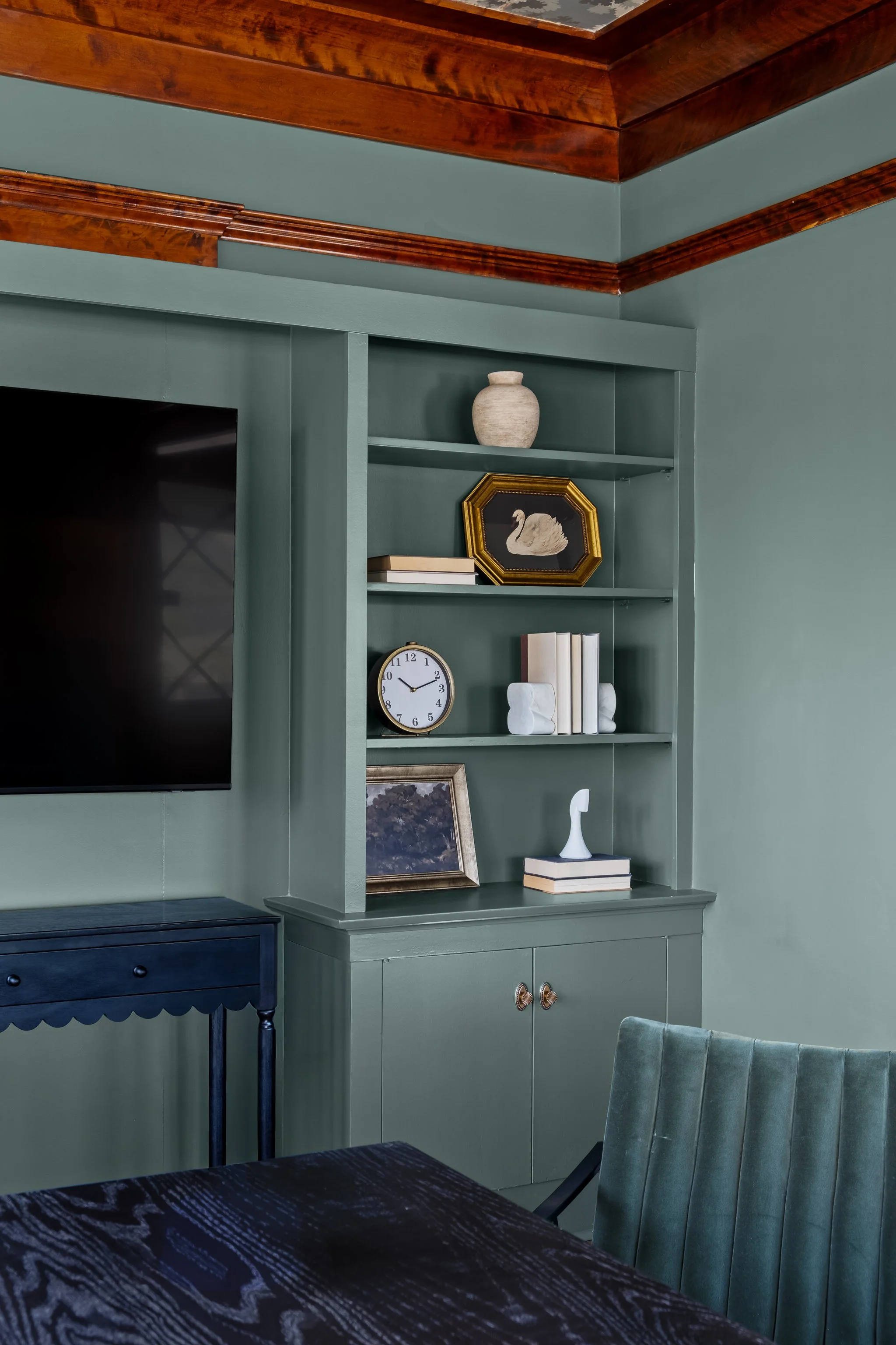 A corner of a living room with a built-in bookshelf painted in green. The shelves hold decorative items including a beige vase, a framed picture of a white swan, stacked books, and a small clock. A dark-colored side table with a scalloped edge is par