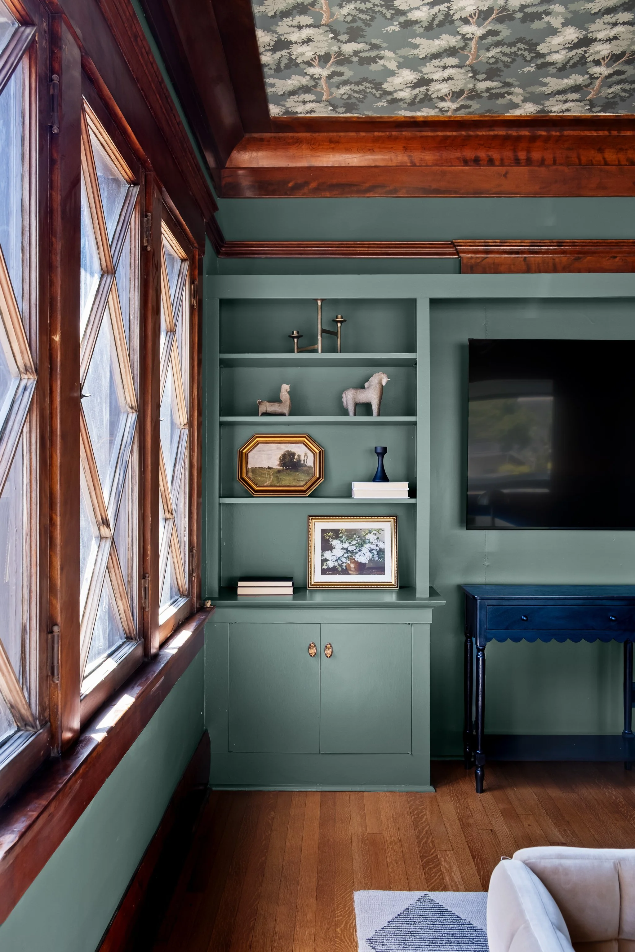 A corner of a living room with a green wall, a built-in green cabinet with decorative items on the shelves, a large flat-screen TV mounted on the wall, a small dark blue table with a scalloped edge, and a beige armchair with a patterned rug on hardwo