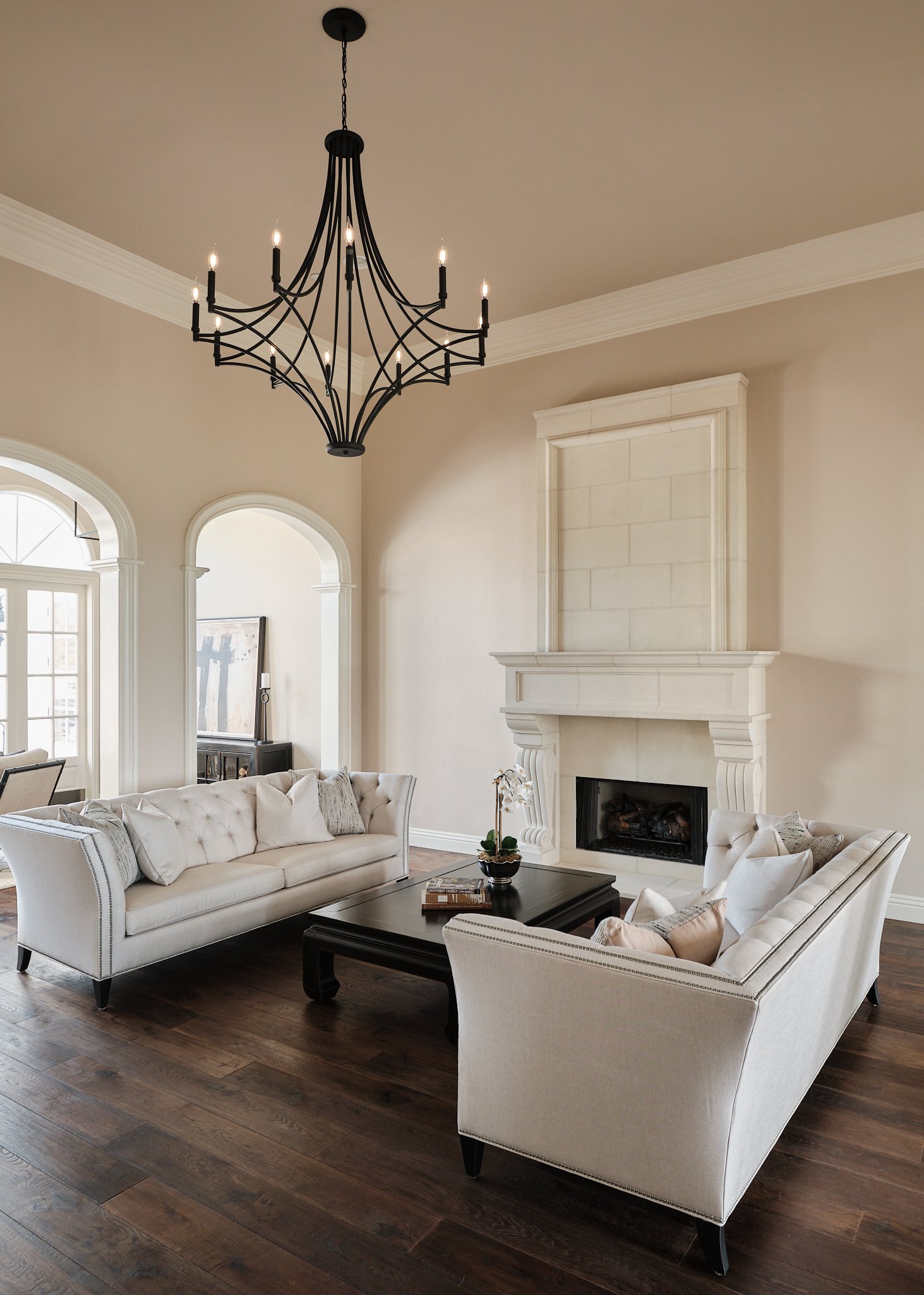 Elegant living room with two white tufted sofas, a black coffee table, a fireplace, and a black chandelier hanging from the ceiling.