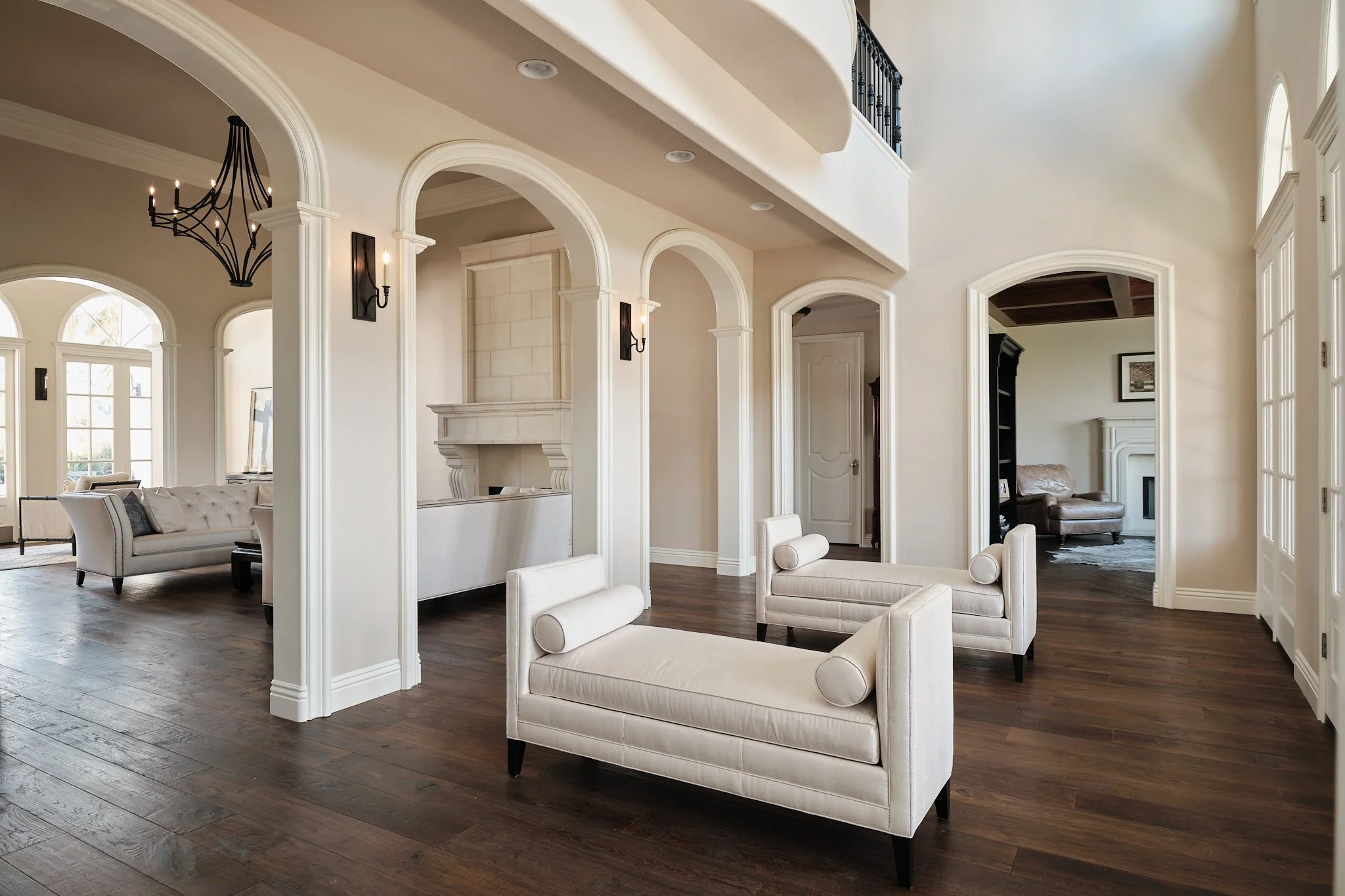 Interior view of a spacious living room with white walls, dark wood flooring, and large arched windows. The room features white upholstered chairs, a beige sofa, and a fireplace with a stone mantel. A modern black chandelier hangs from the ceiling, a