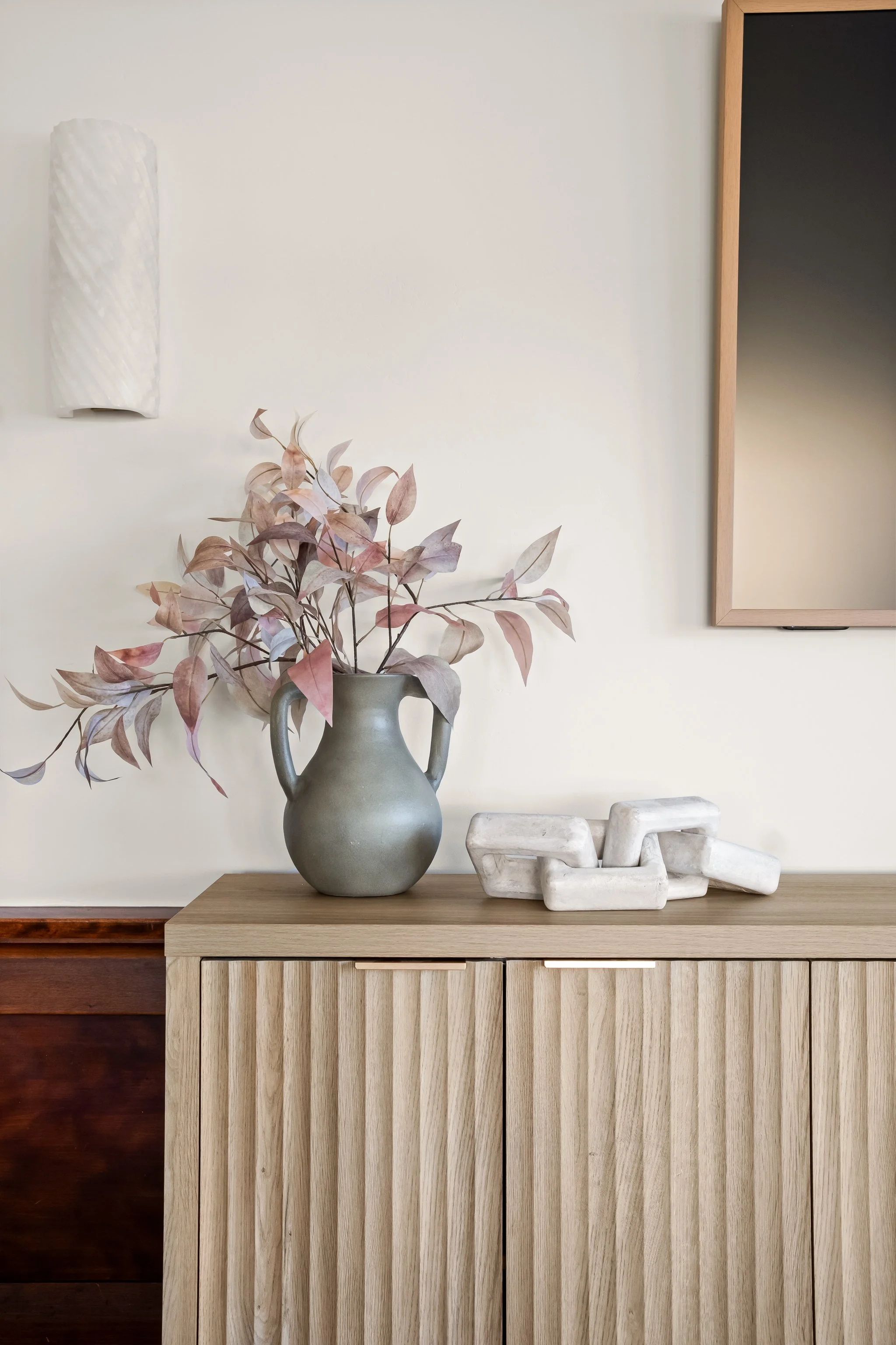A wooden sideboard with a ceramic vase holding dried pinkish-brown leaves, a white abstract sculpture, and a marble sculpture on top. A wall-mounted white textured vase and a rectangular mirror with a wooden frame are in the background.