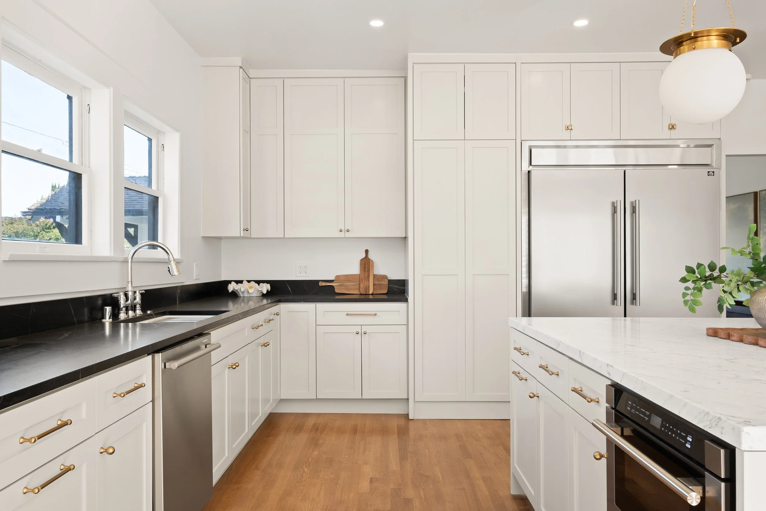 A modern kitchen with white cabinets, black countertops, wooden flooring, stainless steel appliances, and a large window with a view of neighboring houses and trees.