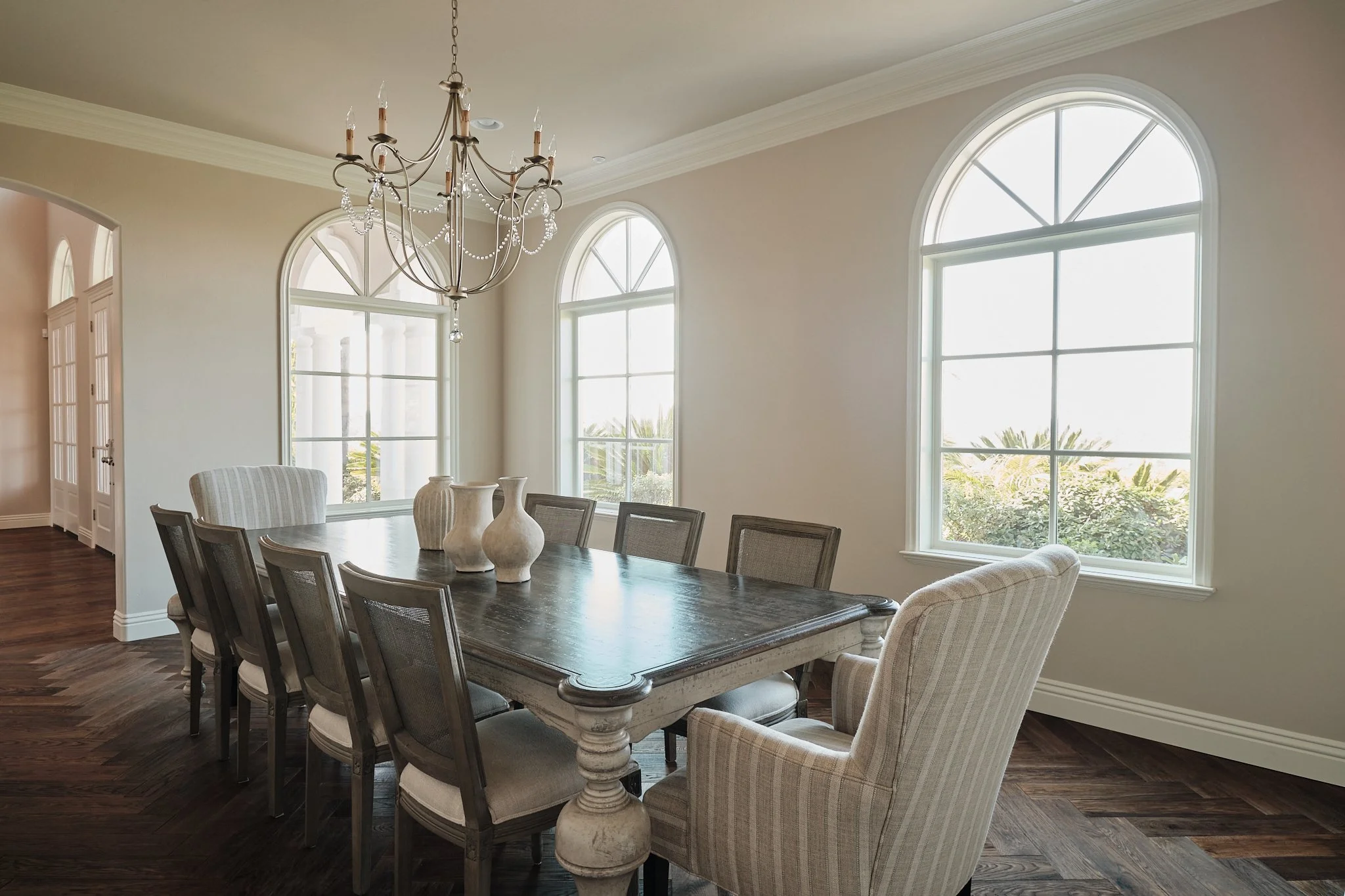A dining room with a large wooden table, eight chairs, a chandelier, and large arched windows letting in natural light.