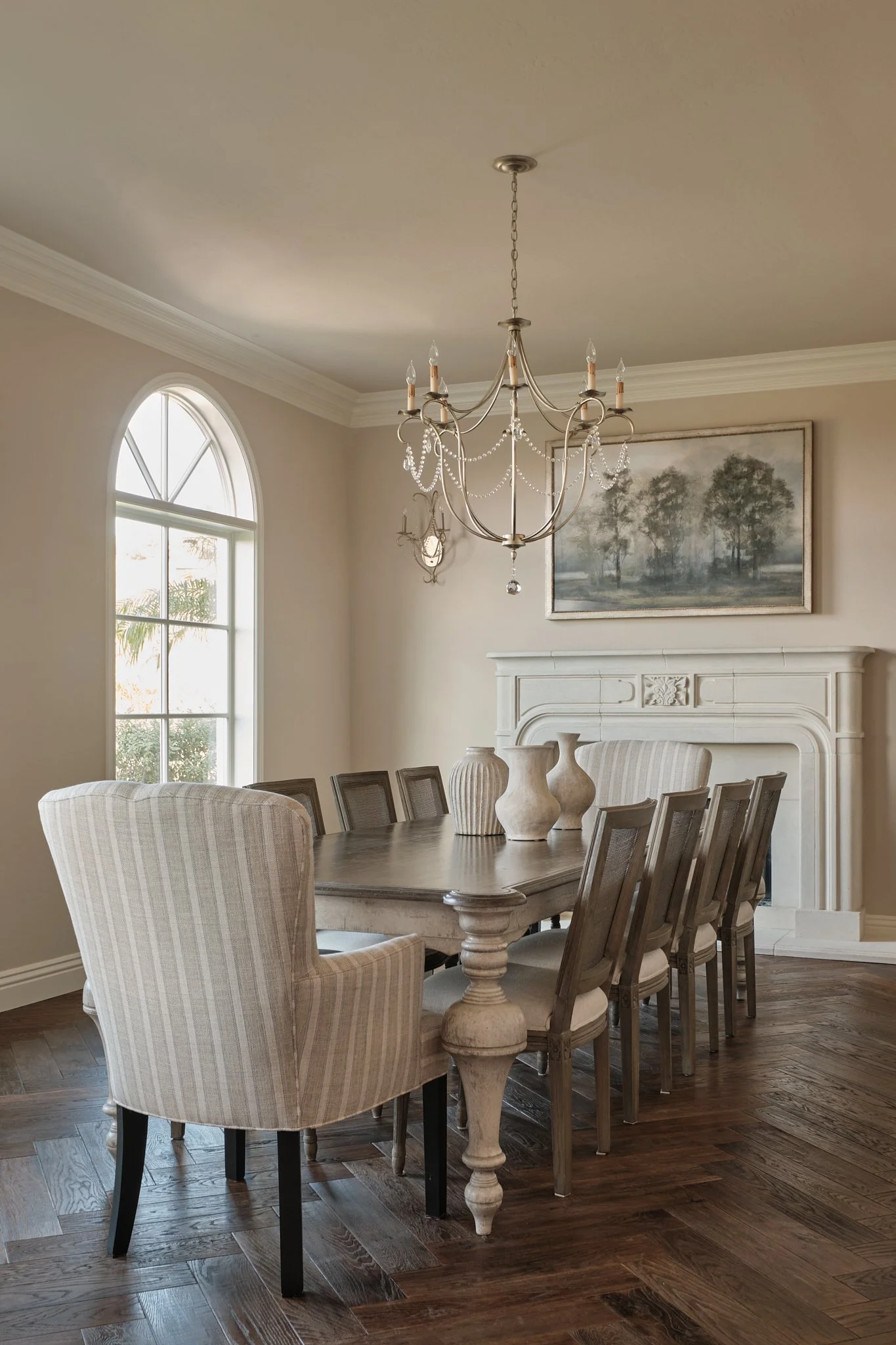 Elegant dining room with a large wooden table, upholstered chairs, a chandelier hanging from the ceiling, and a painting of trees on the wall.