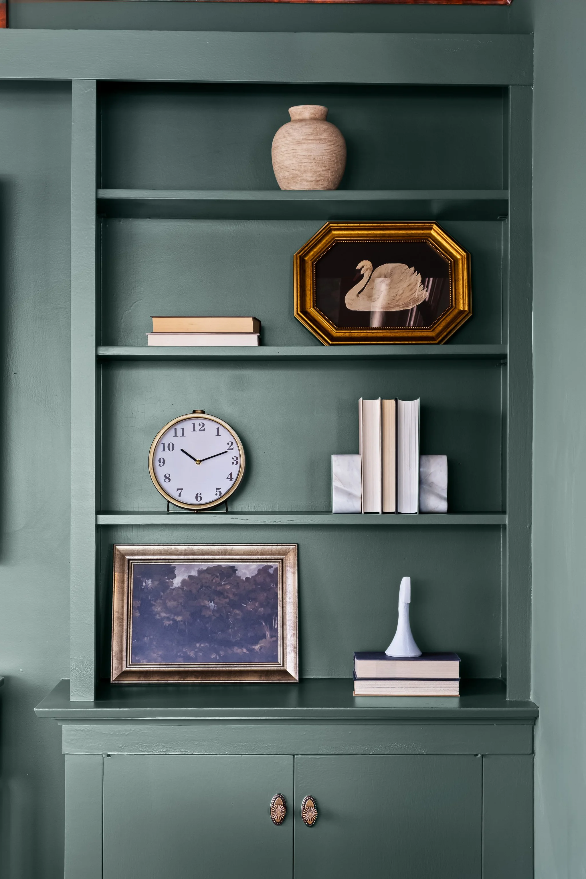 Green built-in bookshelf with decorative items: a beige vase on top, a framed picture of a swan, a clock, a small stack of books, and a framed landscape painting.