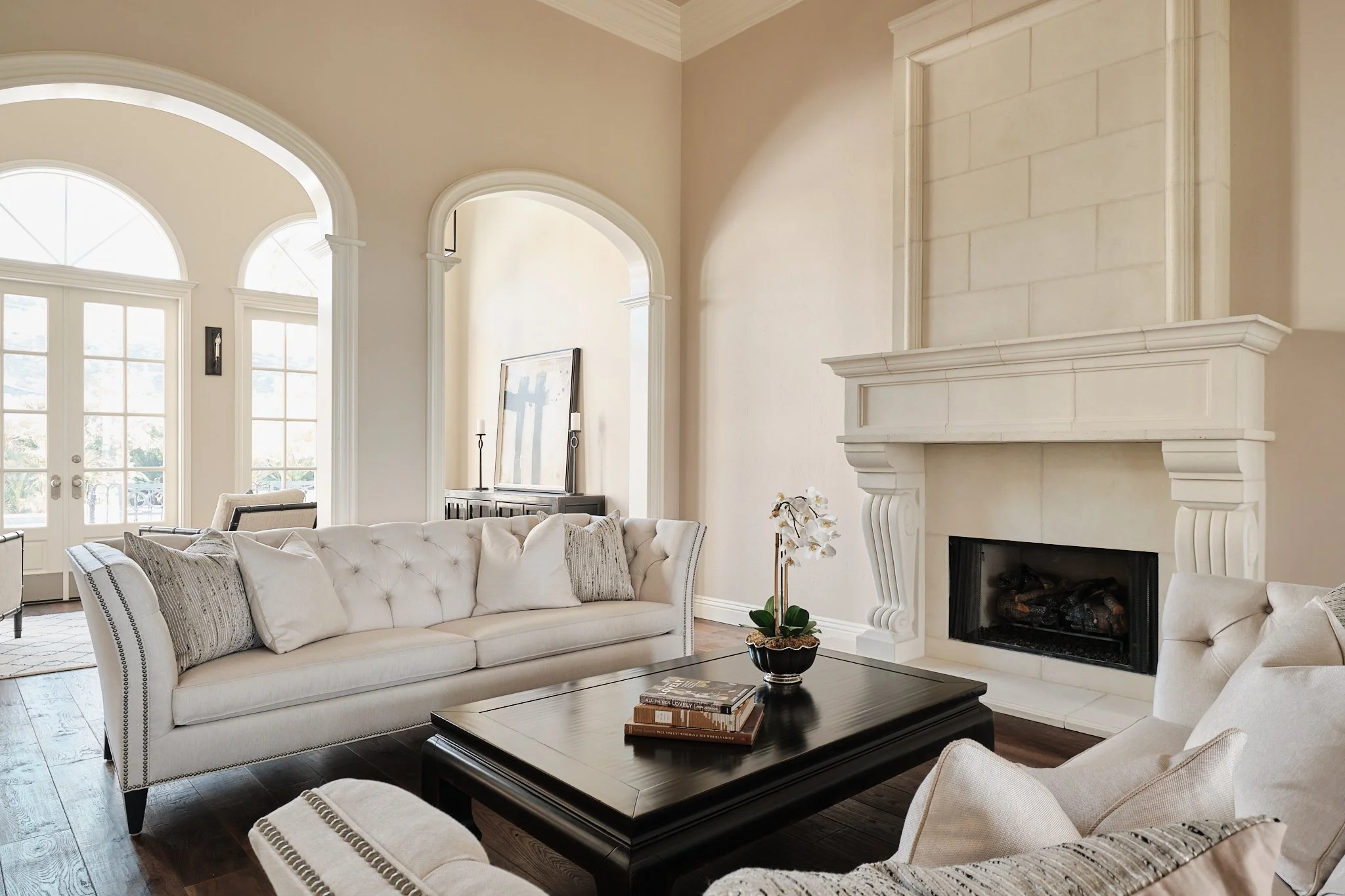 Living room with large windows, white sofas, a black coffee table with books, a fireplace, and an arched doorway with sunlight streaming in.