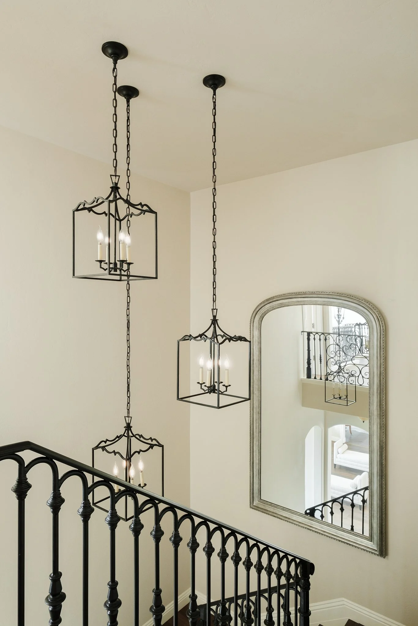 Interior view of a staircase with black metal railing, two black metal chandeliers hanging from the ceiling, and a large mirror on the wall reflecting a balcony with black iron railings and a white outdoor couch.