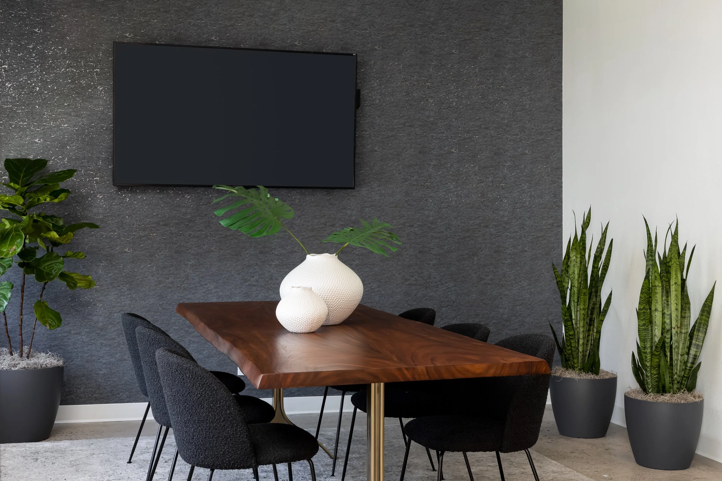 Modern conference room with a wooden table, black chairs, large potted plants, a black wall-mounted TV, and a textured gray wall.