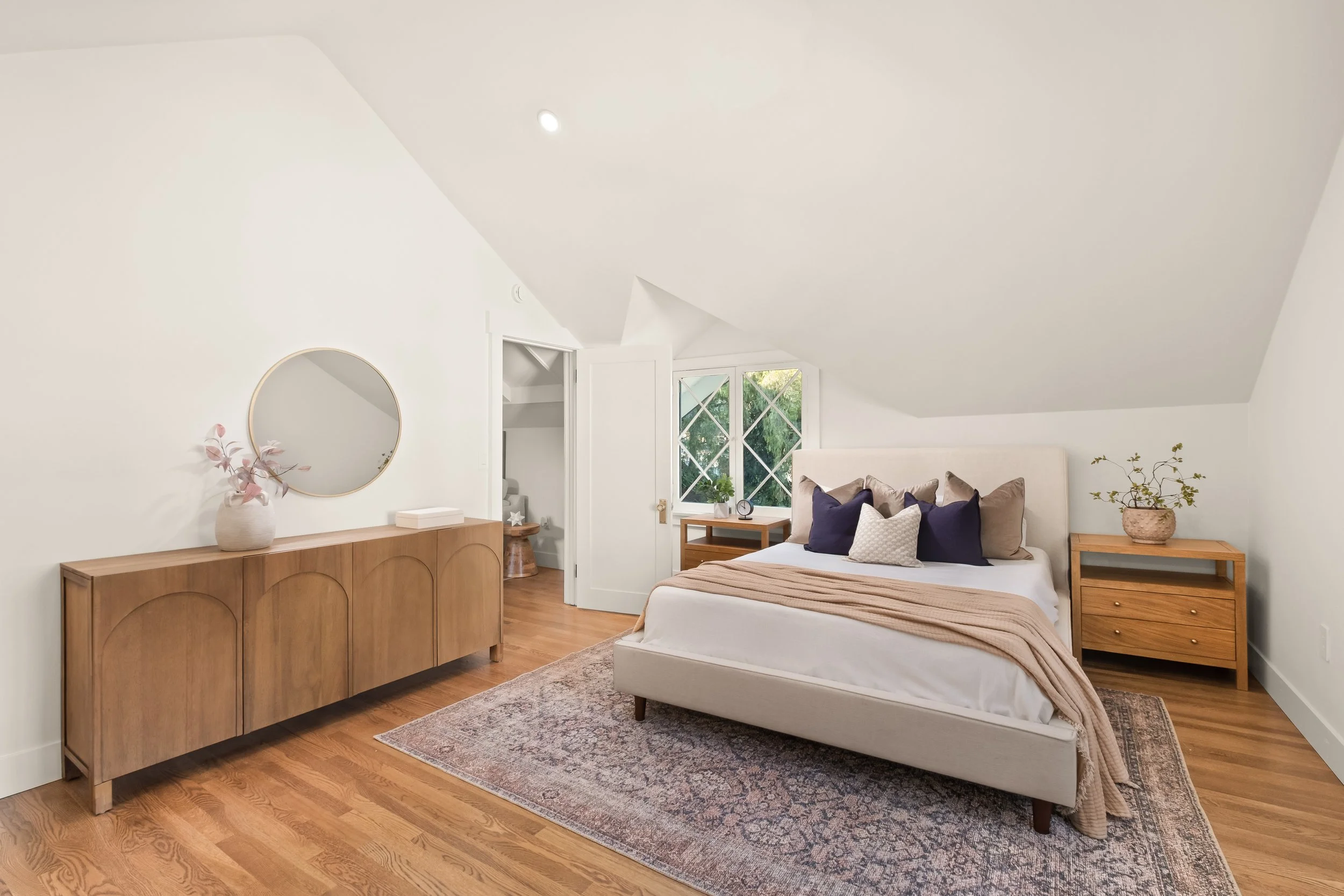 A bedroom with a white bed, beige bedding, and navy and white pillows. Wooden side tables with plants and a clock, a wooden dresser with a mirror and a vase with pink flowers, and a window with a view of greenery.