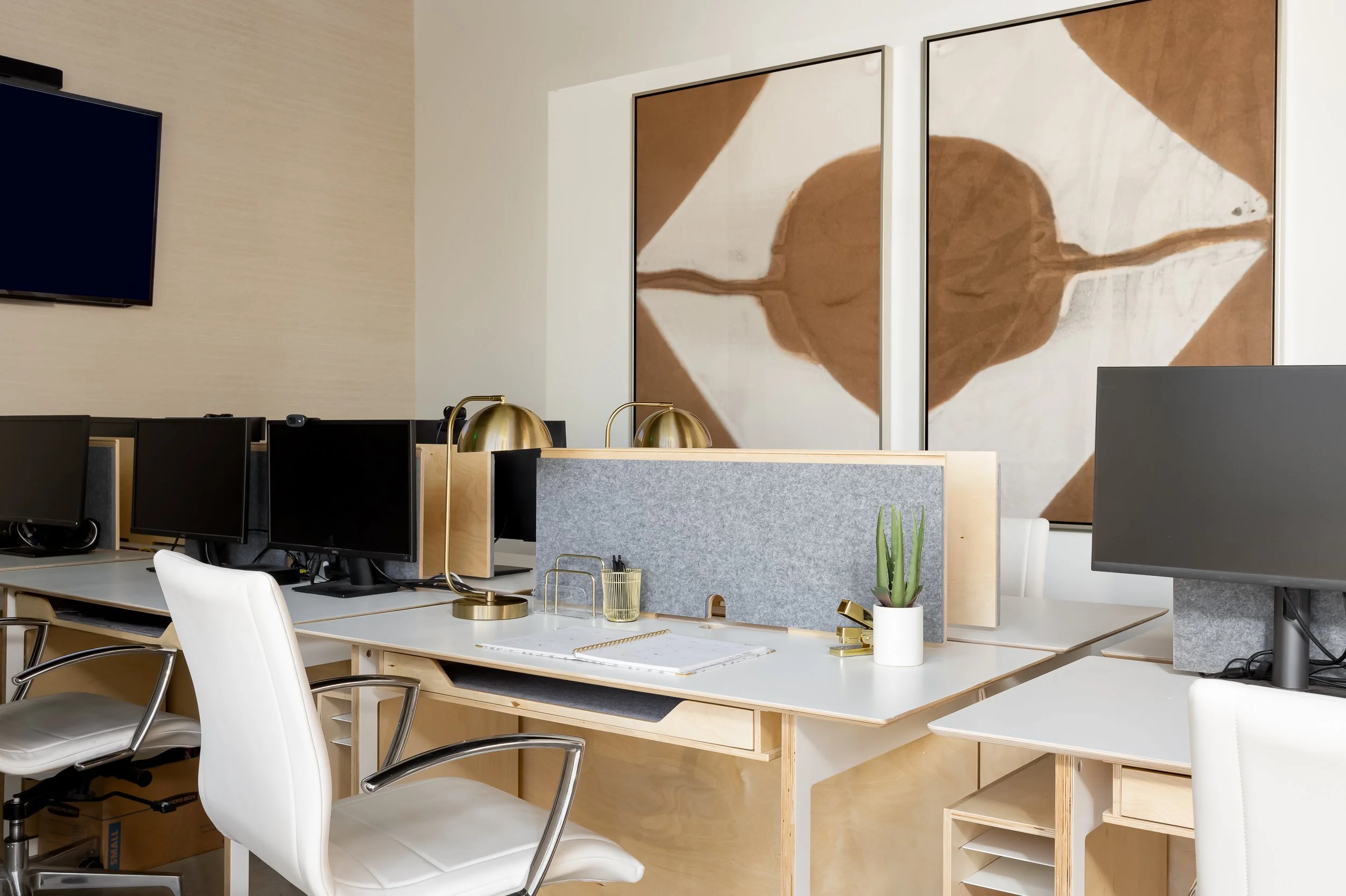 Modern office workspace with multiple computer monitors, a white chair, a wooden desk with a gray felt divider, gold desk lamps, a potted cactus, and a large abstract artwork on the wall with brown and white shapes.