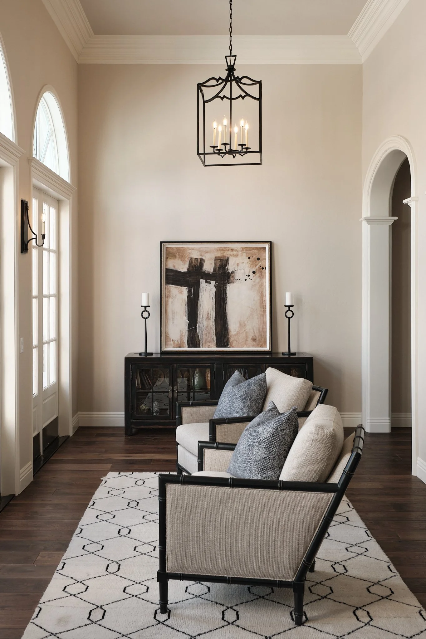 Living room with beige walls, dark wooden flooring, a white patterned rug, and a set of cream-colored armchairs with black frames and gray pillows. A black cabinet holds art pieces and two candleholders with white candles flanking a large abstract bl