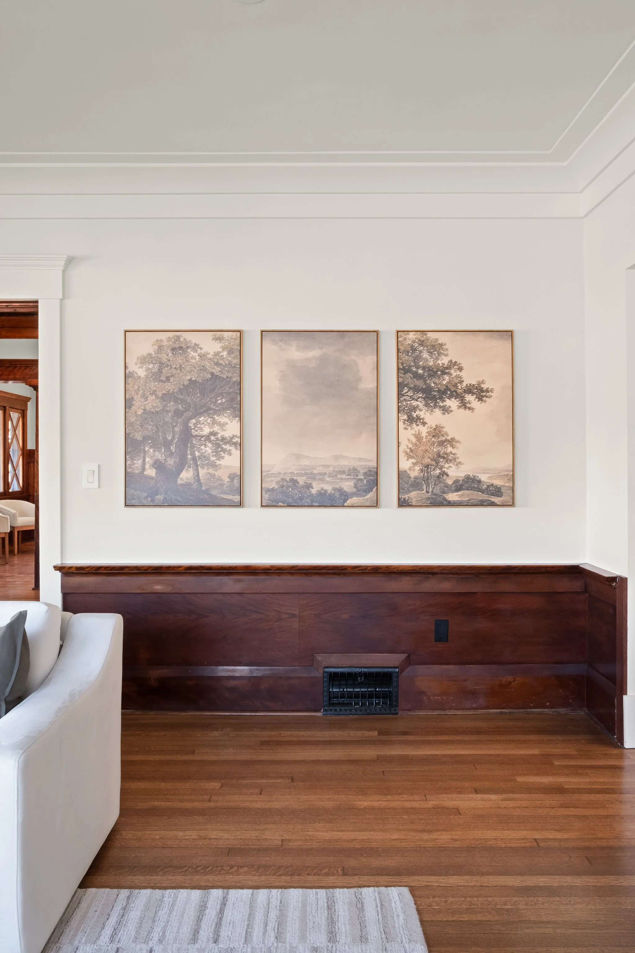 Living room with white walls, three landscape paintings of trees and a landscape, a wooden lower wall panel, a vent, hardwood floor, and part of a white sofa and light-colored rug.