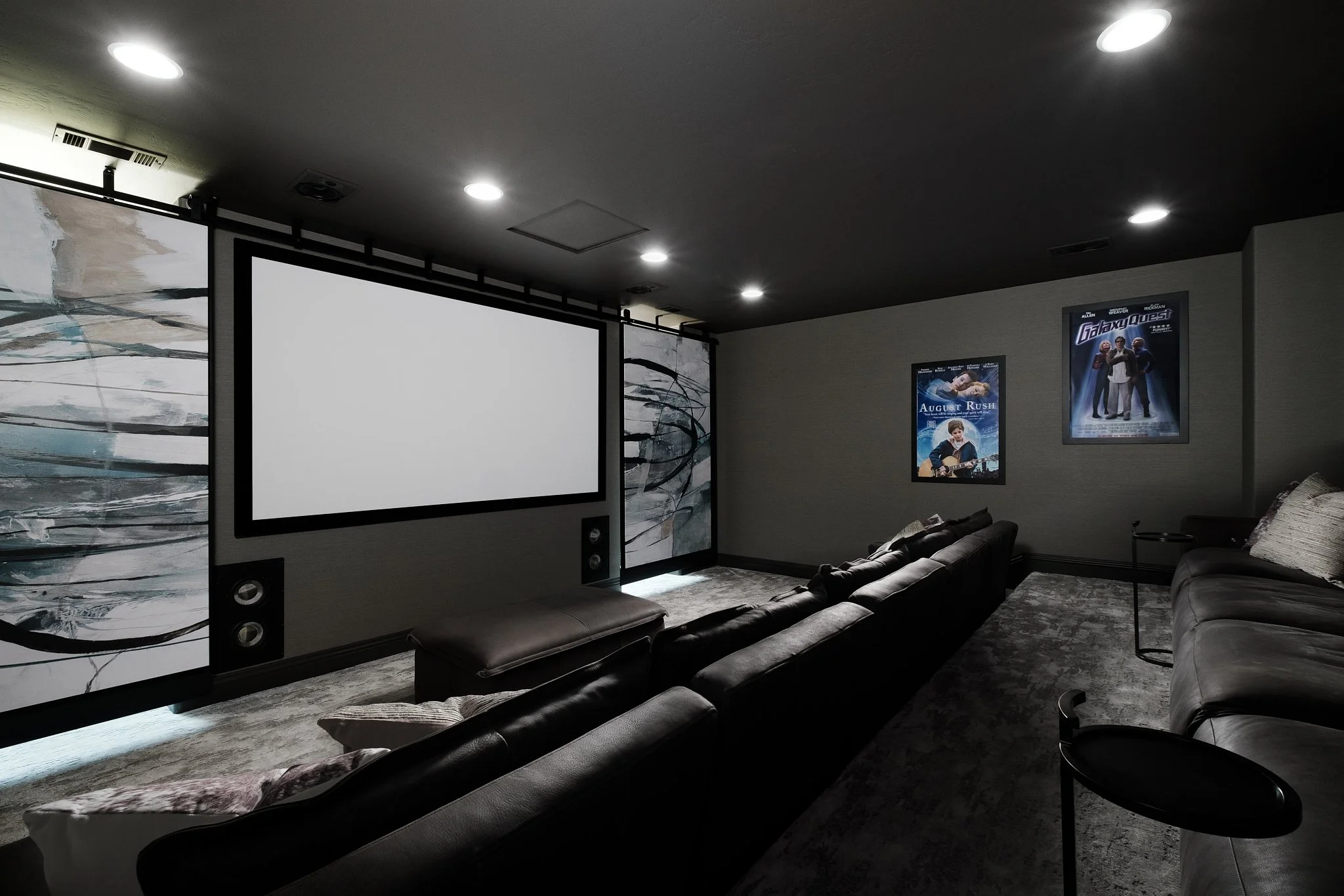 A home theater room with a large white projection screen, black border, and movie posters on the wall, including 'Galaxy Quest' and 'August Rush'; black couches arranged for viewing, with side tables and ambient lighting.