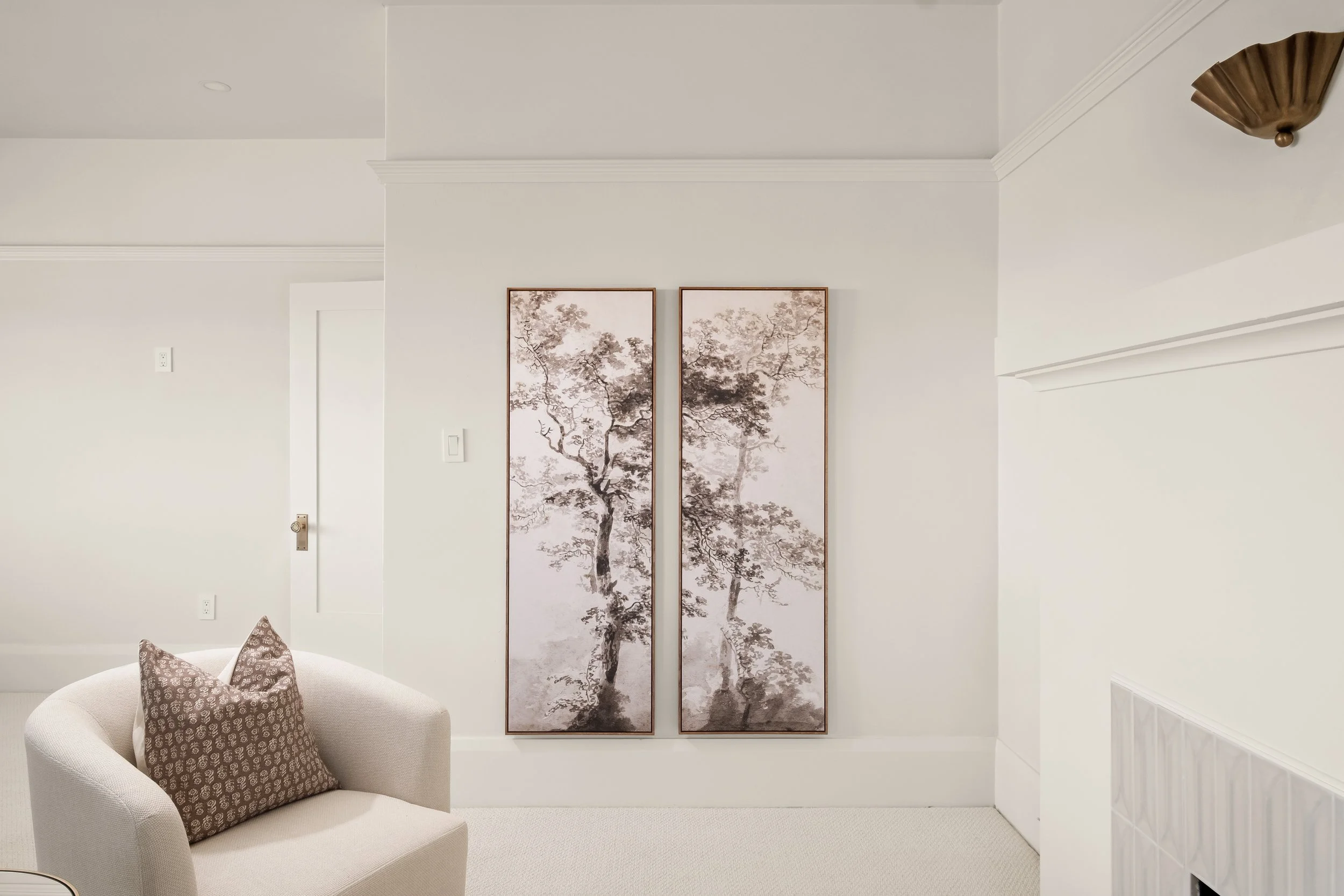 Living room corner with a white curved sofa holding decorative pillows, a two-panel wall art of trees, white walls, crown molding, and a ceiling light fixture.