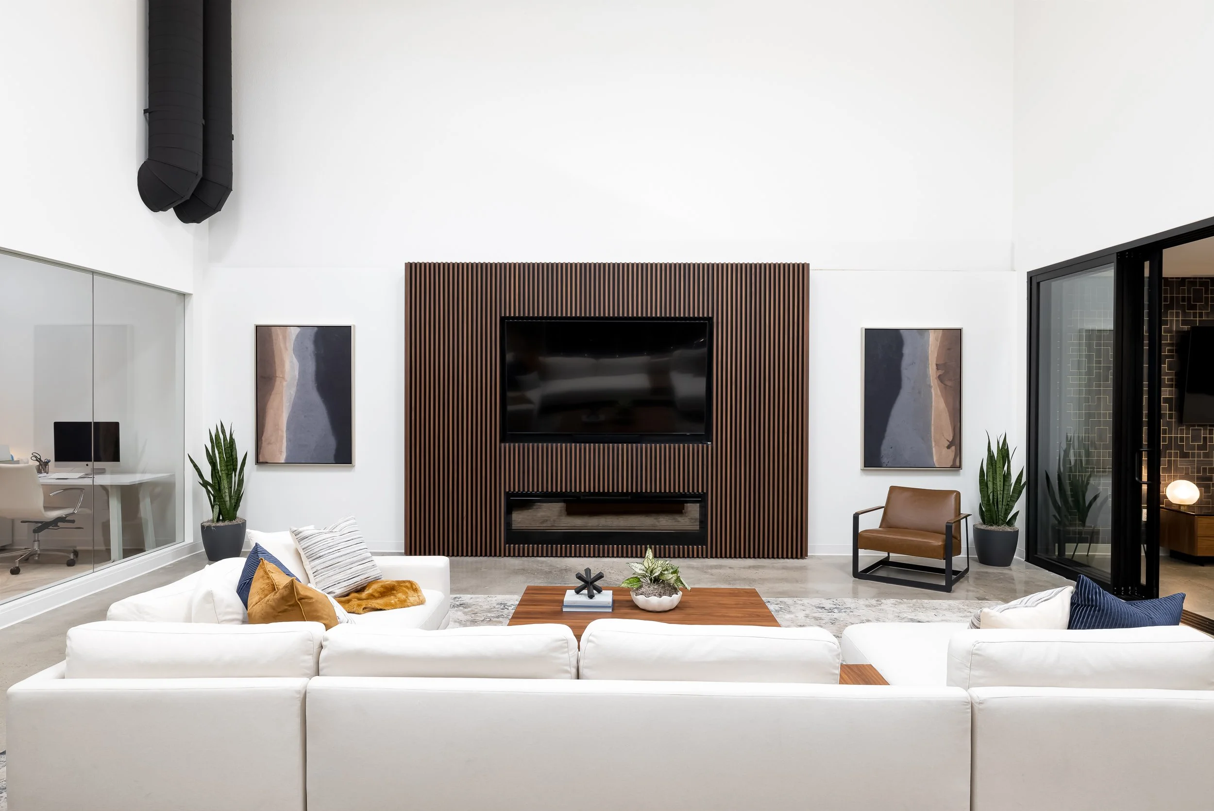 Modern living room with a white sofa, decorative pillows, wooden coffee table, large black-framed television, two artwork pieces, indoor plants, and a brown armchair against a white wall with a fireplace.