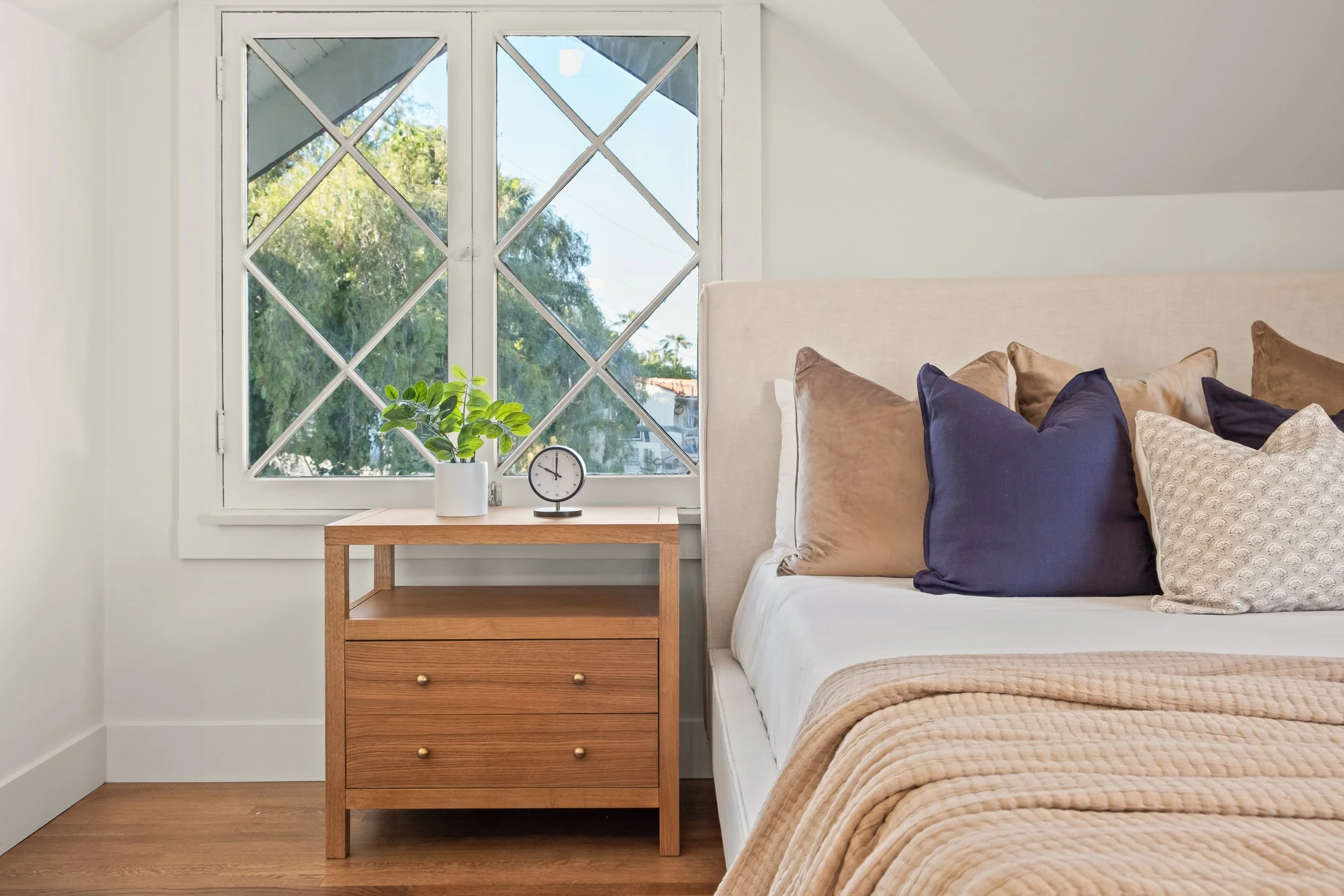 Bedroom with a beige headboard, beige and navy pillows, a wooden nightstand with gold knobs, a white pot with green plant, and a small clock, next to a window with a view of trees.