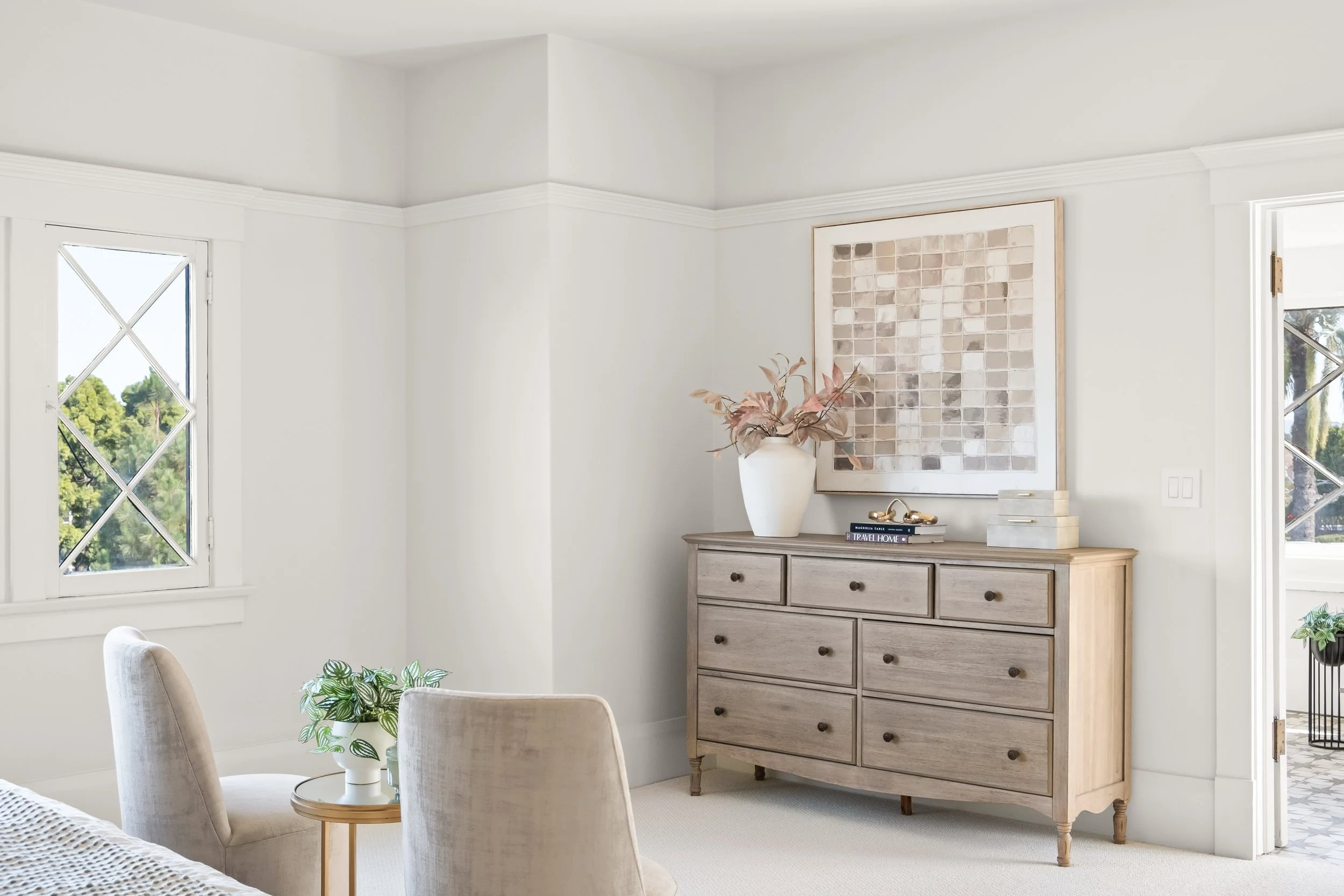 A room with white walls and ceiling, featuring two windows with diamond-shaped grid pattern, a wooden sideboard, a framed tile artwork, a white vase with pinkish leaves, and a few decorative books and objects.