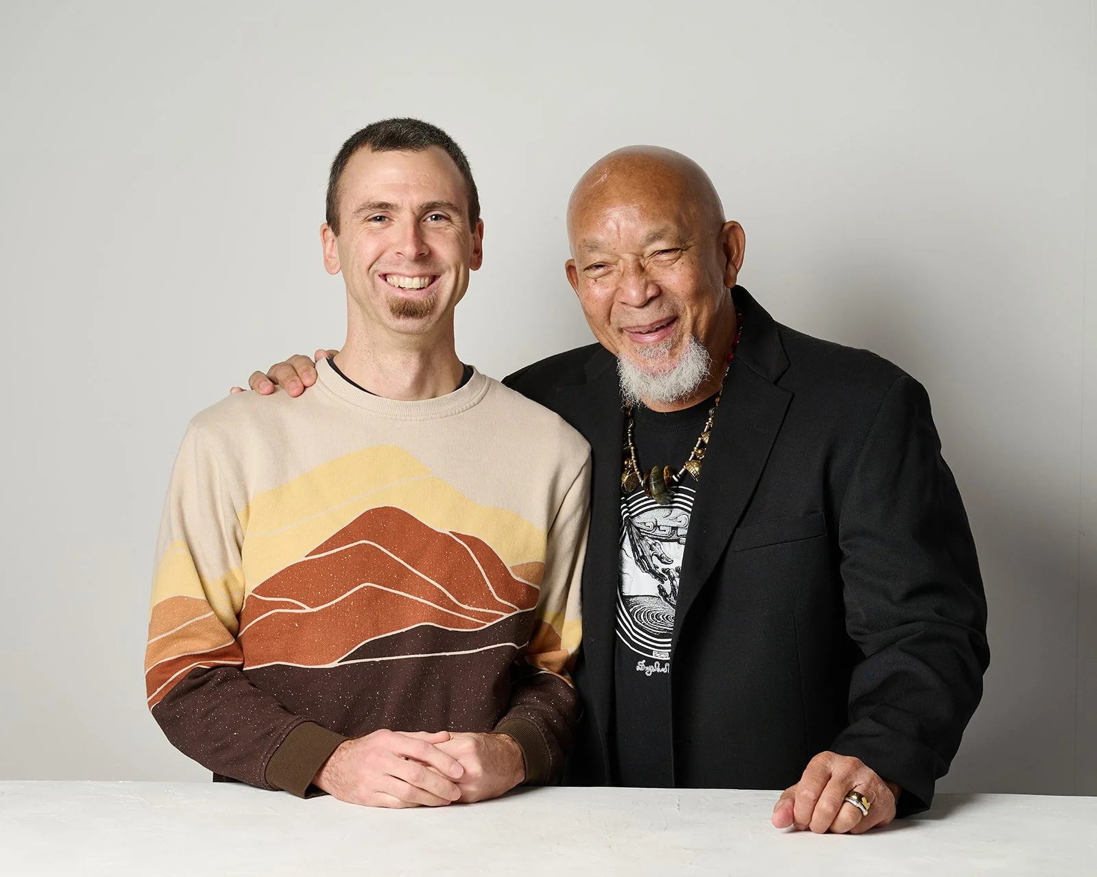 Two men stand together, smiling, with one man placing his hand on the other's shoulder. The man on the left is younger, with short brown hair, wearing a beige sweater with a mountain landscape design. The man on the right is older, with a bald head and white beard, wearing a black blazer over a graphic t-shirt, and an ornate necklace.