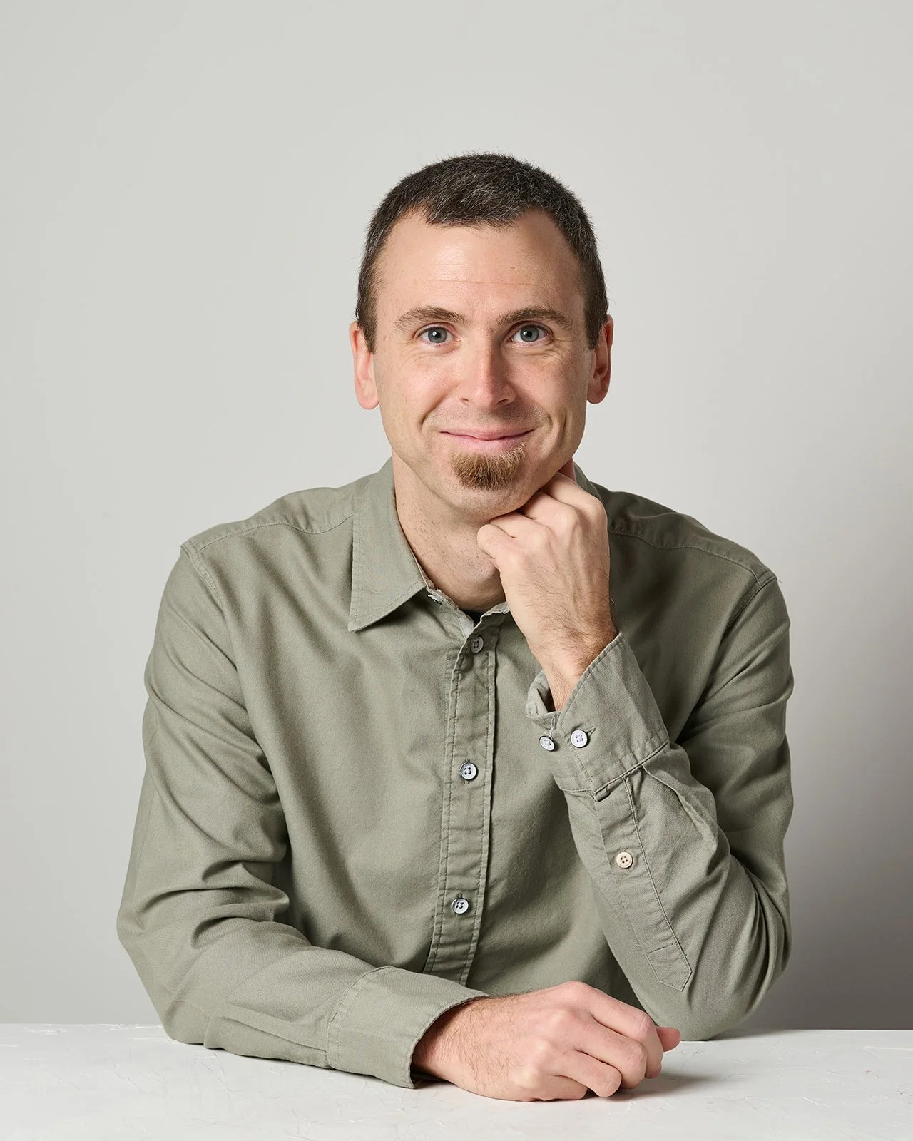 Portrait of a man with short dark hair, a goatee, wearing a light green button-up shirt, smiling, resting his chin on his right hand, sitting at a white surface against a plain light gray background.