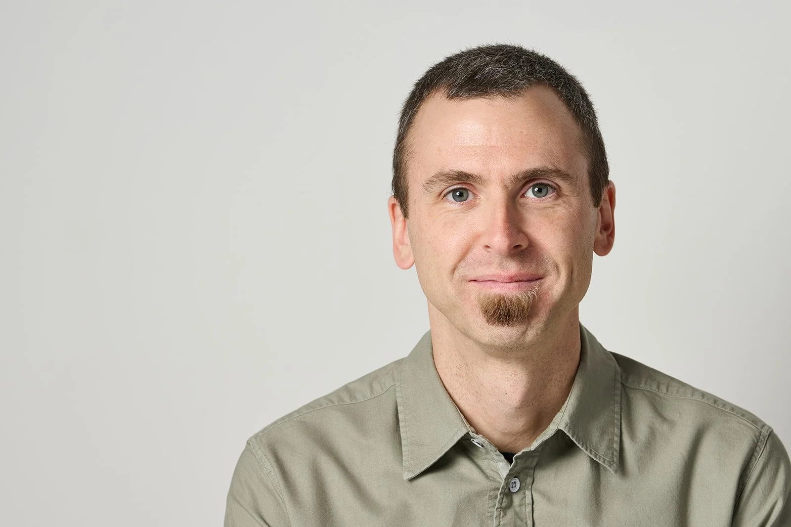A man with short brown hair, a slight goatee, wearing a light gray shirt, smiling against a plain light gray background.
