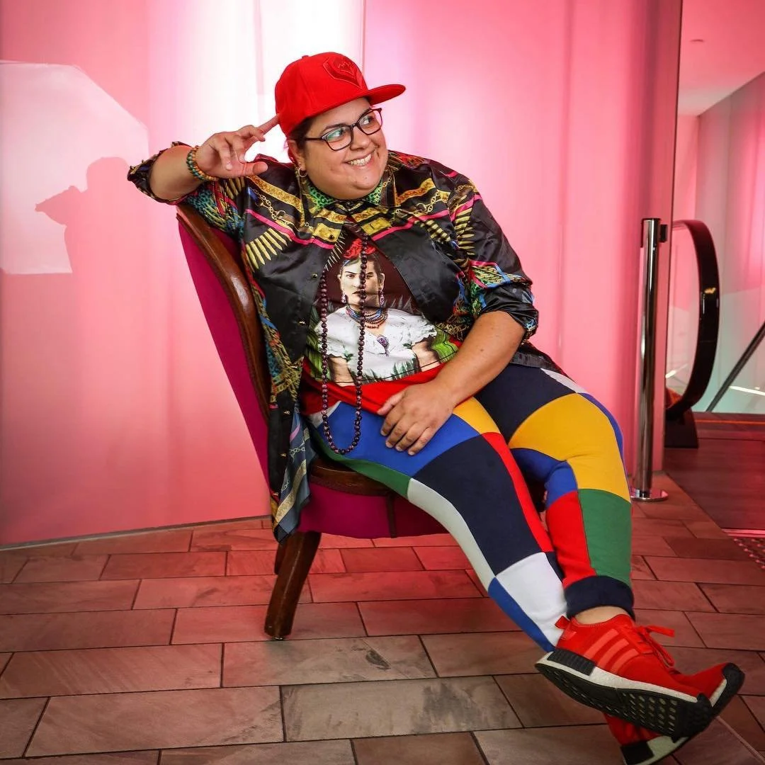 Mz Rizk sits on a chair in brightly colour clothing, smiling