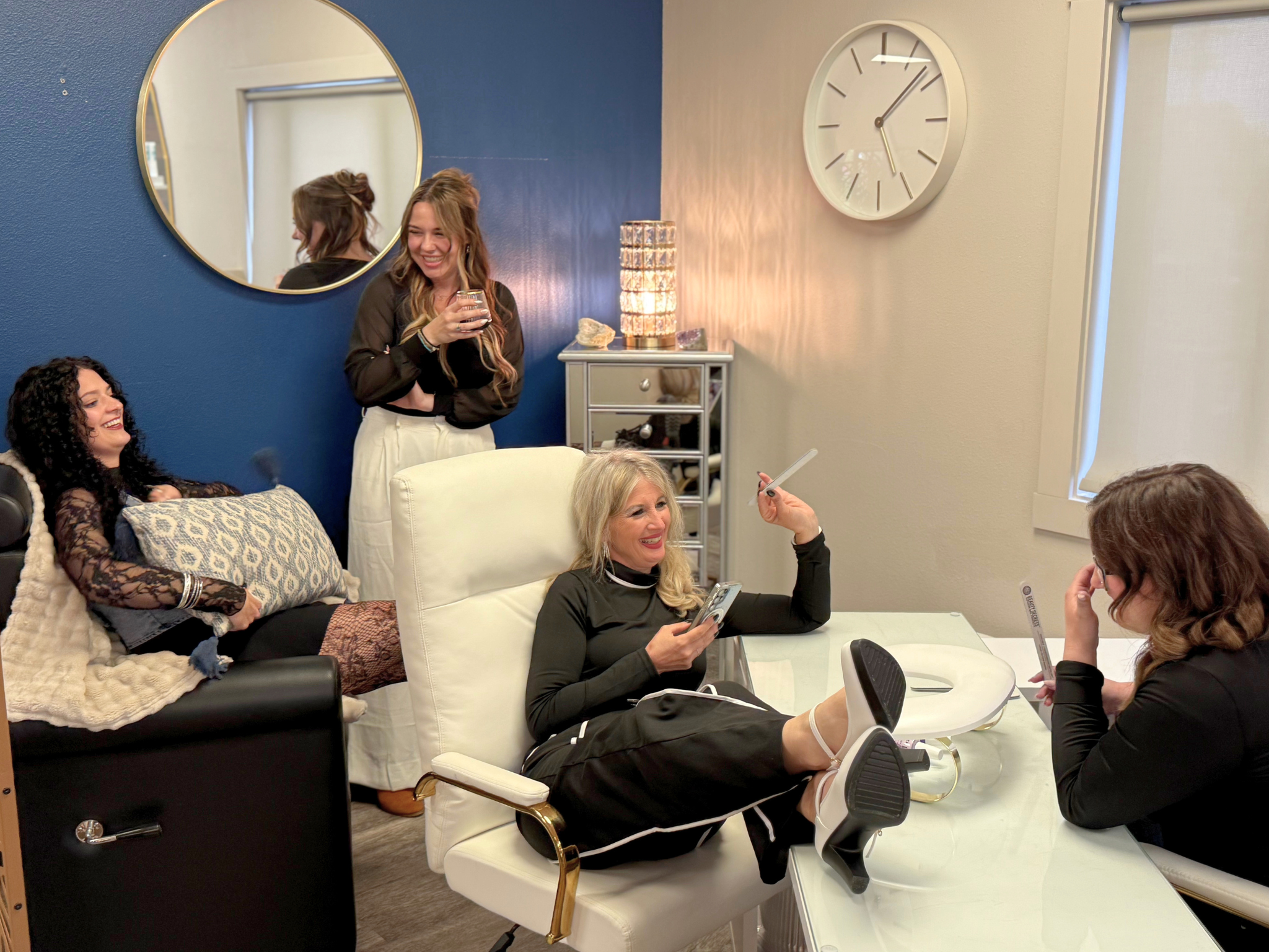 A group of women in a living room having a conversation. One woman is sitting on a white armchair holding a phone, and others are sitting or standing around her, smiling. The decor includes a blue accent wall, a large mirror, a clock, a table with candles, and a bed with a patterned pillow and blanket.