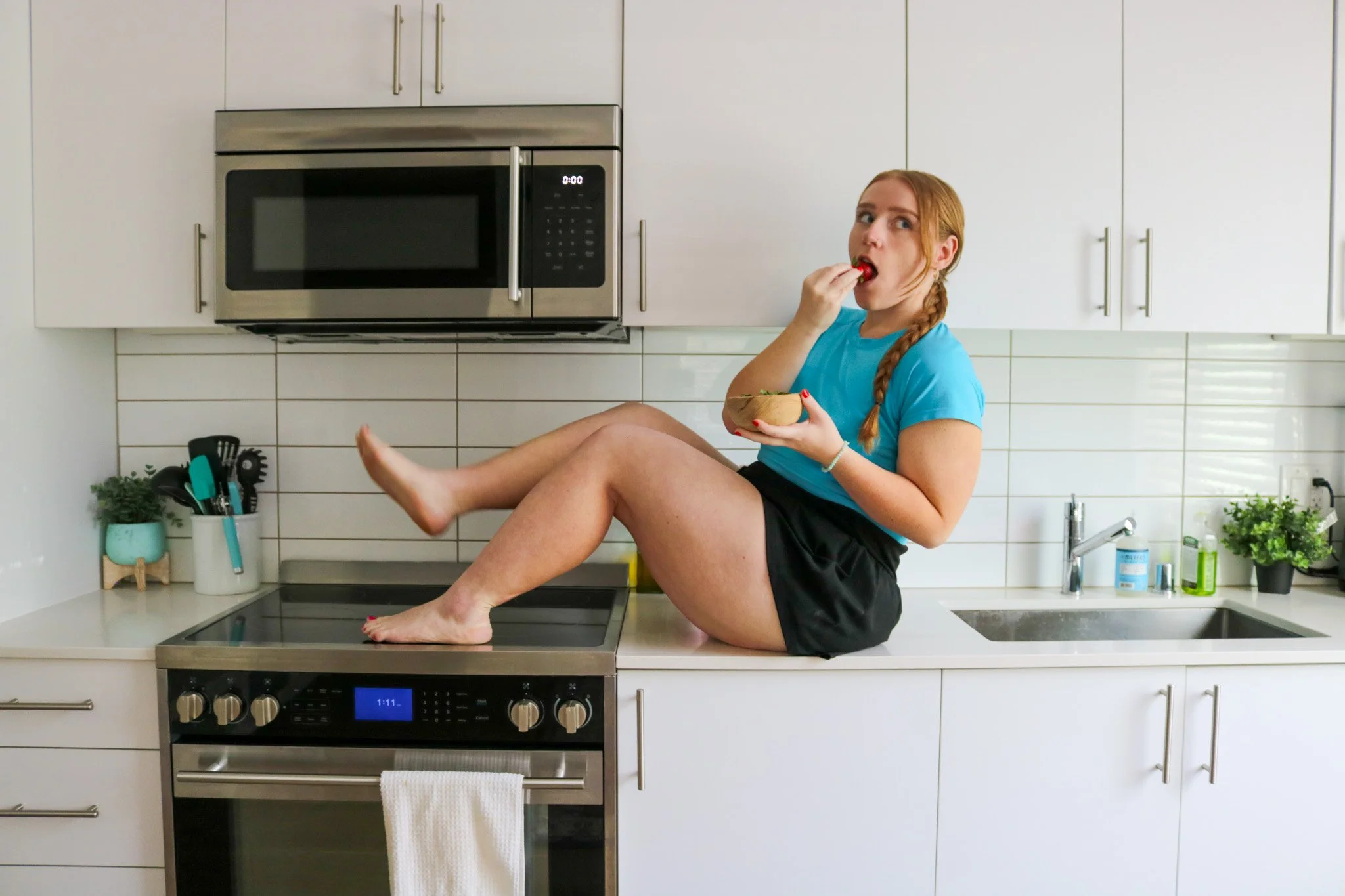 A woman with red hair braided, sitting on a kitchen countertop with her legs up, eating a strawberry and holding a bowl of salad. The kitchen has white cabinets, a microwave, and various kitchen utensils.