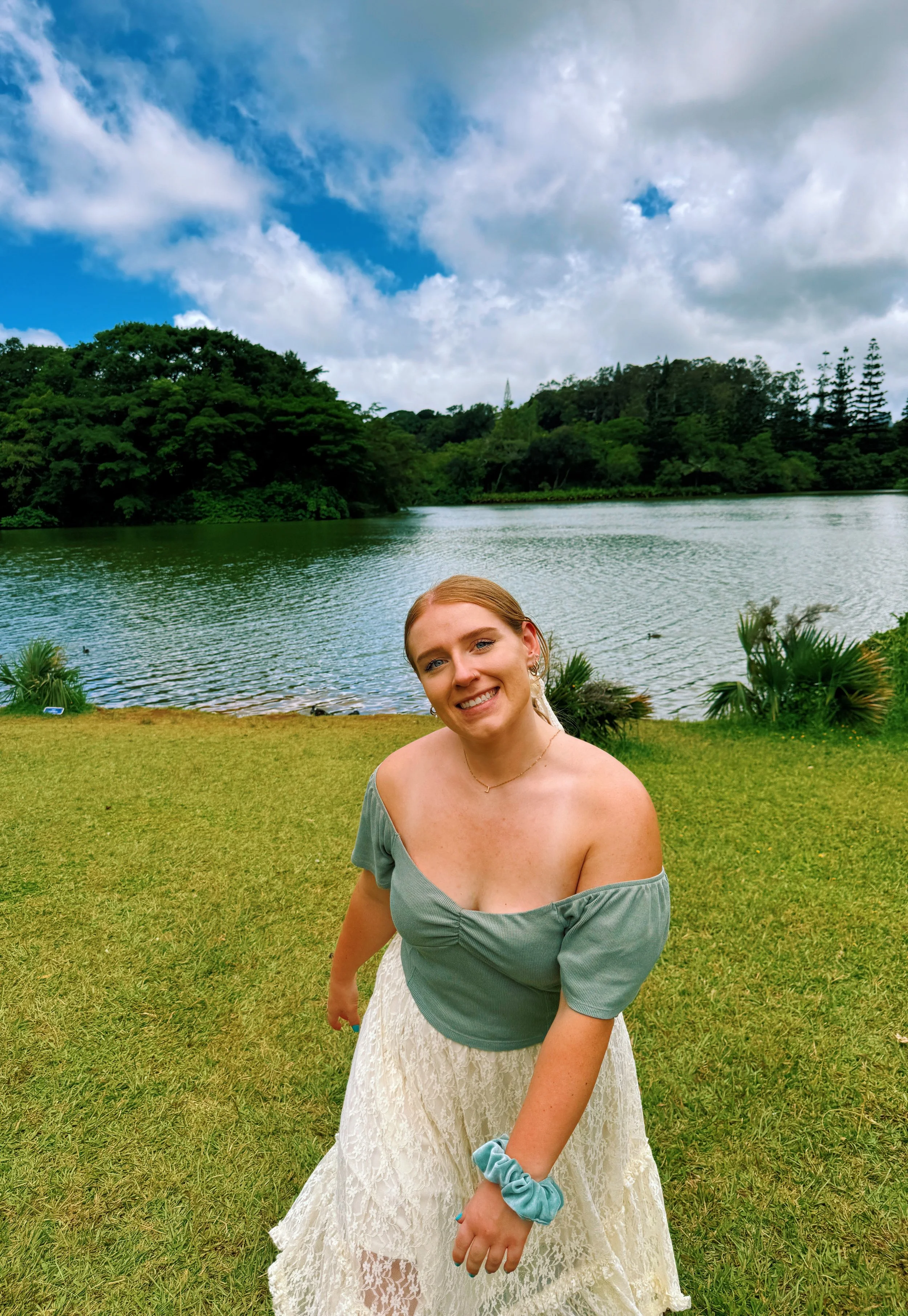 A woman with blonde hair smiling outdoors near a lake with lush green trees and a partly cloudy sky in the background.