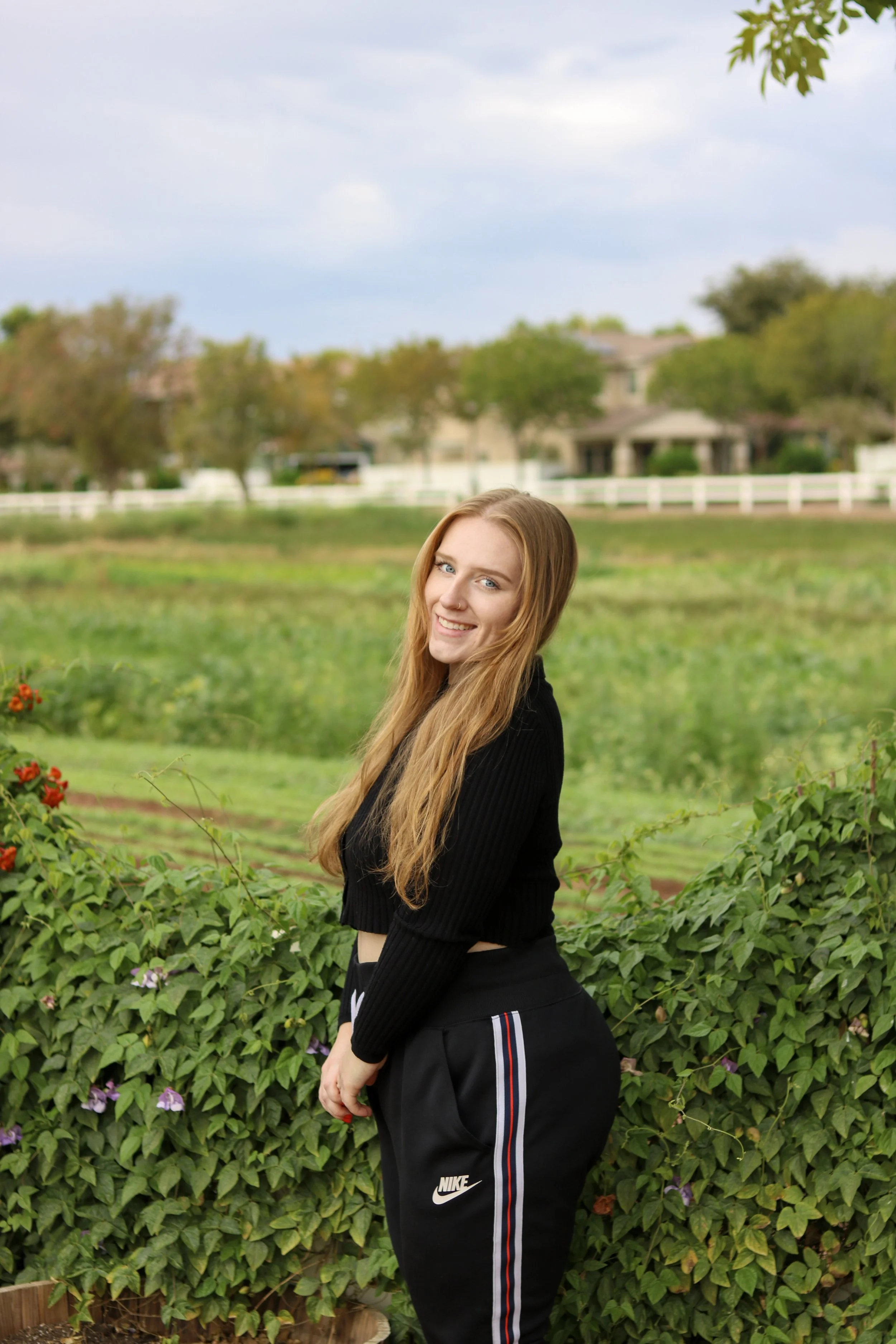 A young woman with long blonde hair and blue eyes wearing a black long-sleeve top and Nike sweatpants standing outdoors in a garden with green bushes and colorful flowers. In the background, there is a grassy area, trees, a house, and a cloudy sky.