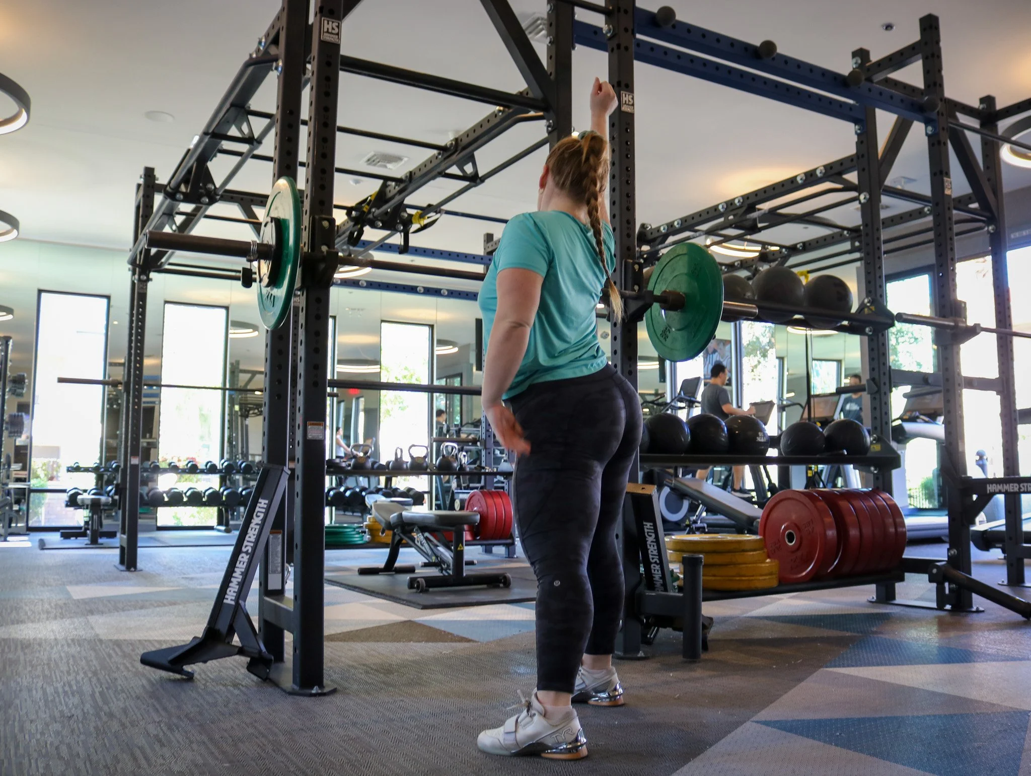 A woman with a braid, wearing a teal t-shirt, black pants, and white sneakers, is lifting a barbell with green weight plates in a modern gym. The gym has large windows, black metal workout equipment, and several other people working out in the background.