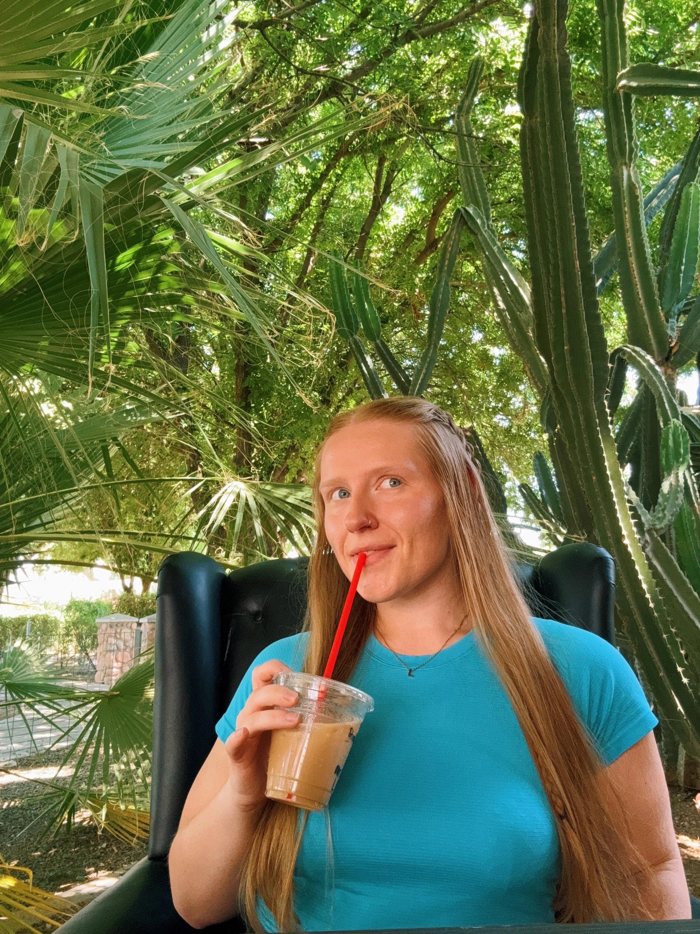 A young woman with long red hair, wearing a blue shirt, is sitting outdoors under green trees and drinking iced coffee through a straw from a clear plastic cup.