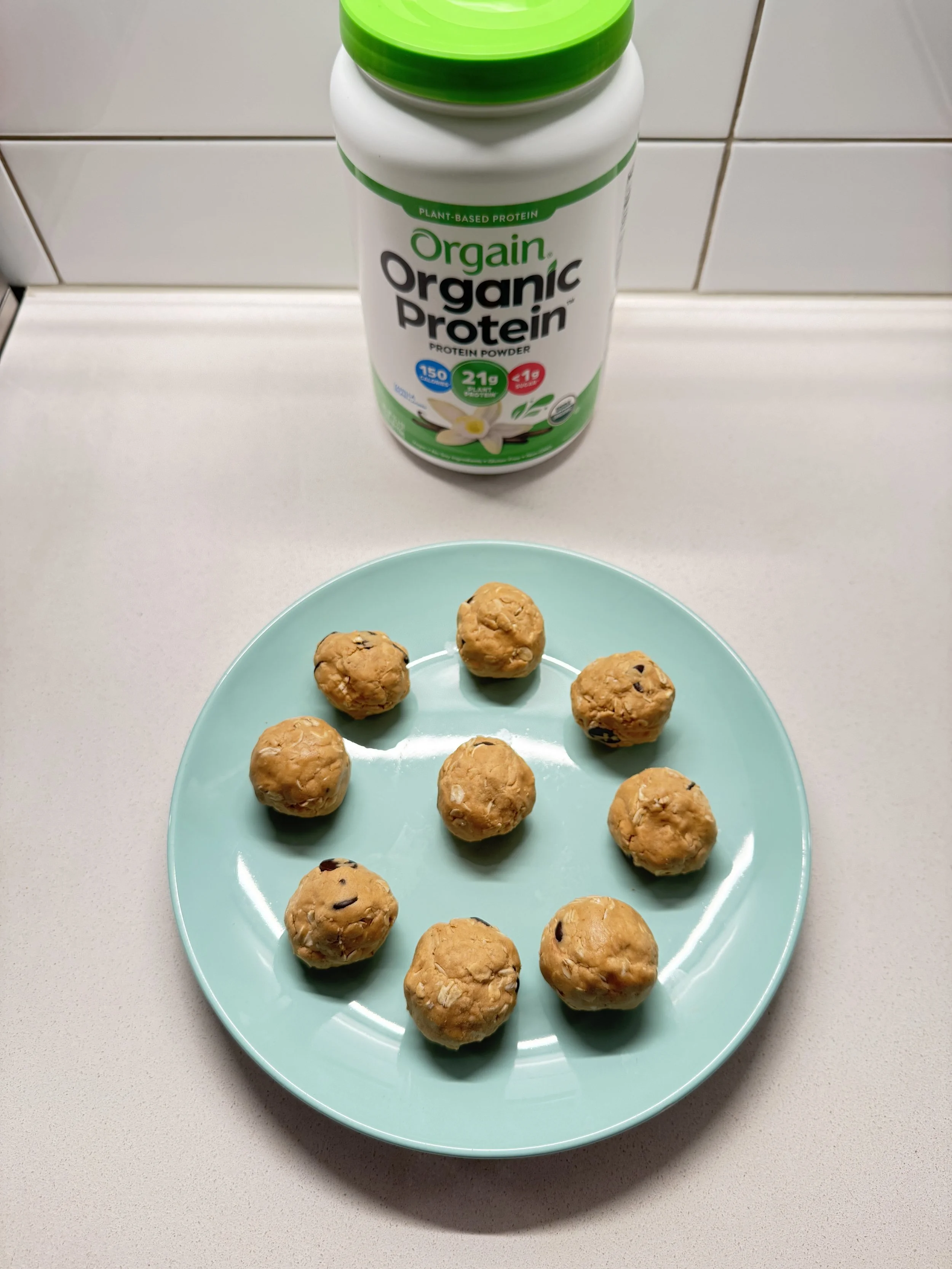 Protein balls on a teal plate sitting on the kitchen counter next to Orgain Organic Protein Powder.