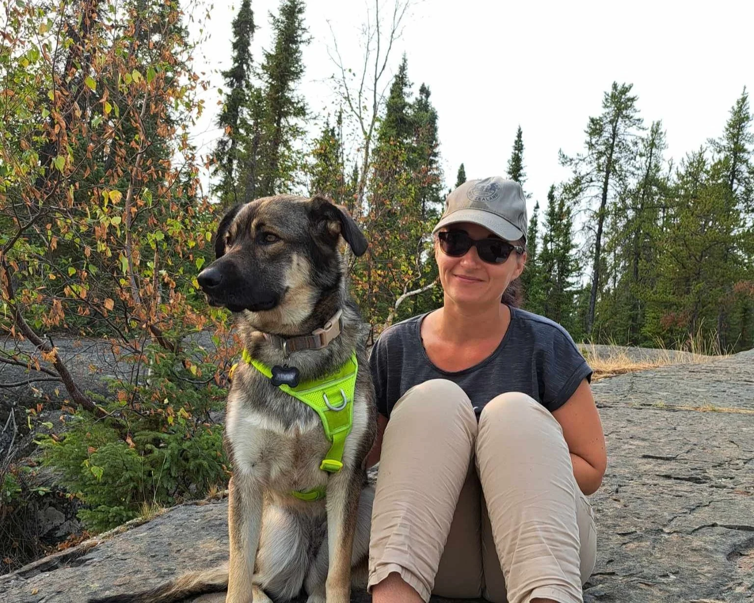 Photo d'une femme et son chien, assis sur des rochers devant des arbres. Le chien (gauche) est un husky/berger allemand moyen et porte un harnais jaune fluo. La femme (droite) porte pantalon beige, t-shirt gris, casquette et lunettes de soleil.