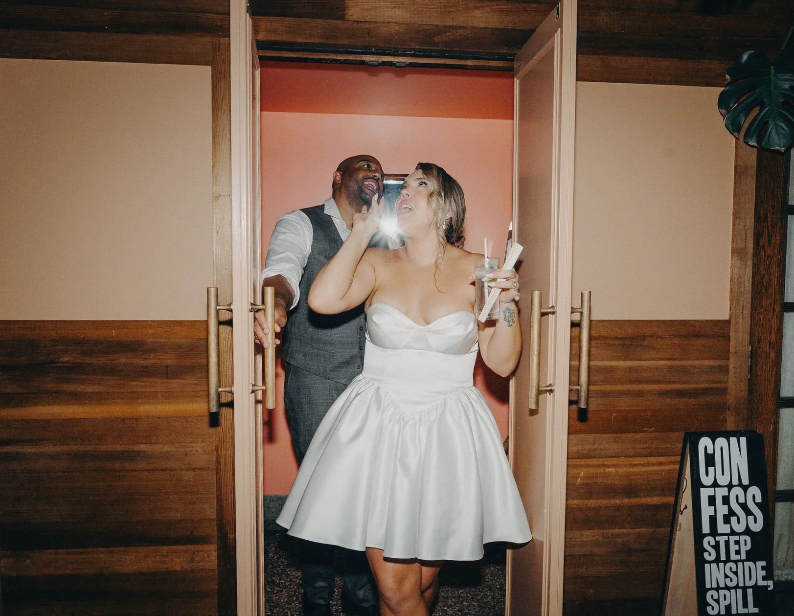 A bride in a strapless white wedding dress and a man in a vest and shirt sharing a moment inside a doorway at a wedding reception, with a sign on the floor reading "Confetti - Step Inside, Spill".