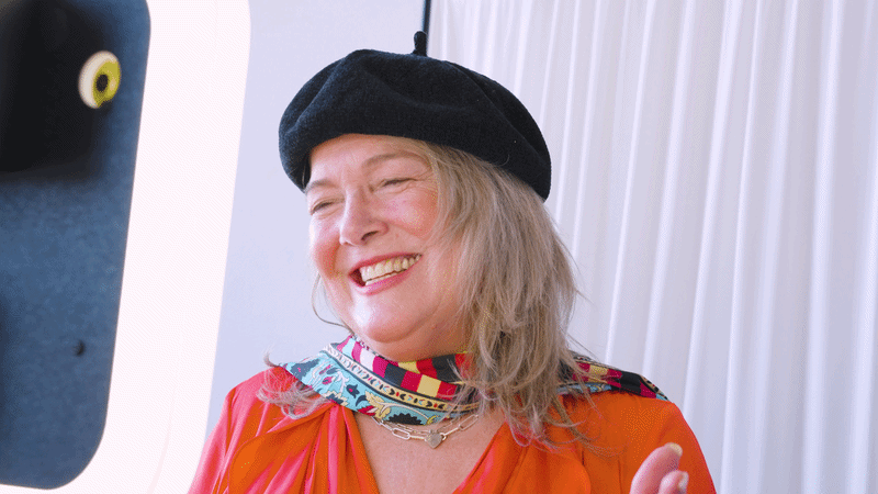 A woman with light-colored hair wearing a black beret, colorful scarf, and an orange top, smiling. A part of a camera is visible in the foreground.