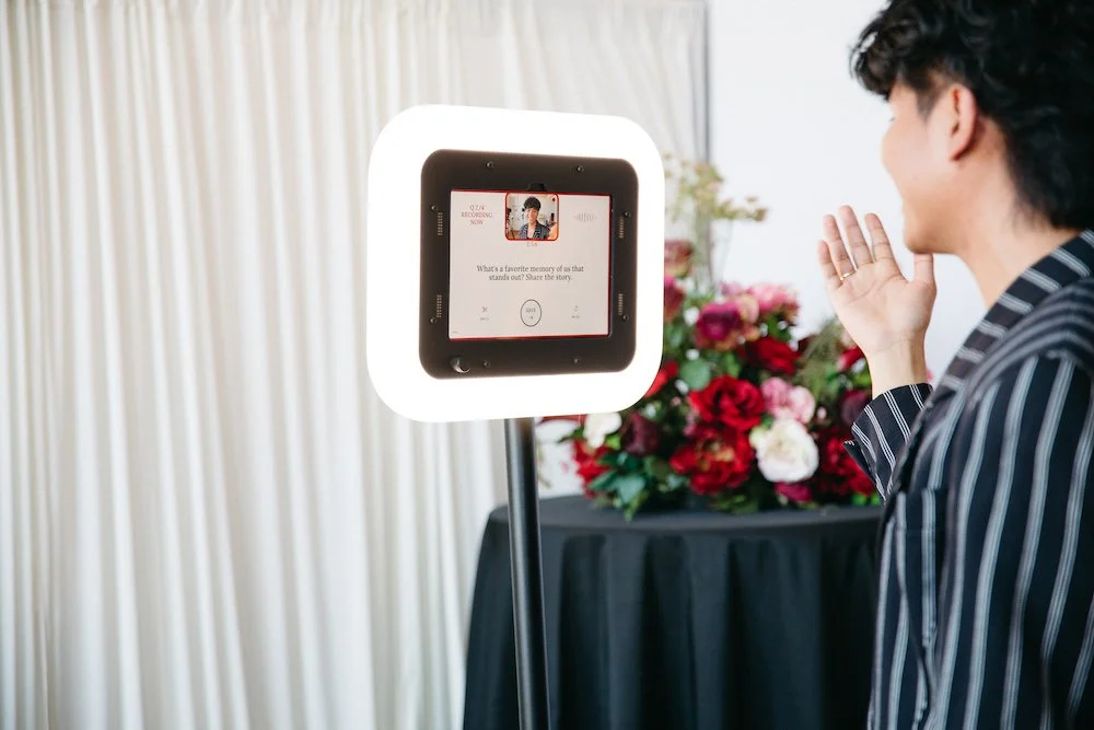 A person is taking a selfie with a device surrounded by a ring light, with a floral arrangement and curtain in the background.