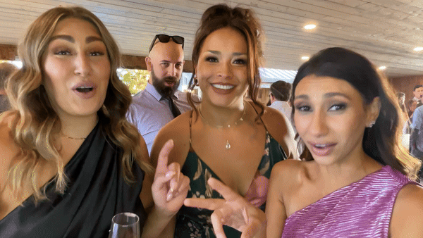 Three women and one man at a social event or party, smiling and engaging with the camera, with a wooden ceiling overhead and other people in the background.