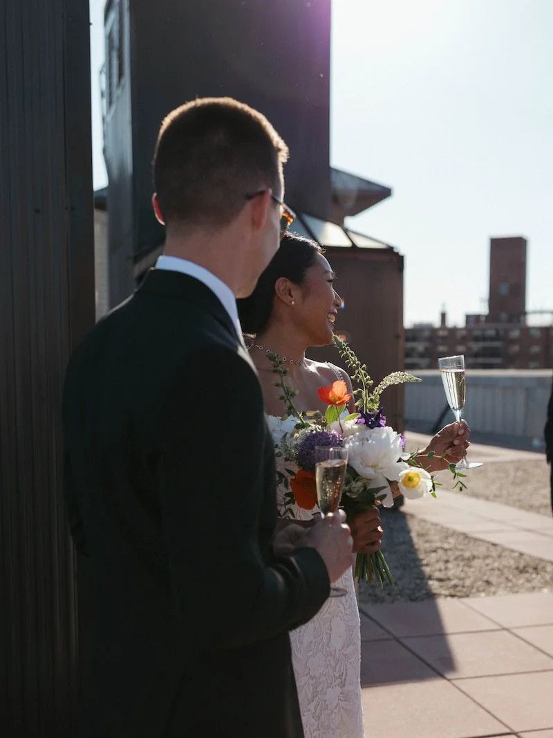 A bride and groom celebrating outdoors, holding champagne glasses with the bride holding a bouquet of flowers, both smiling.